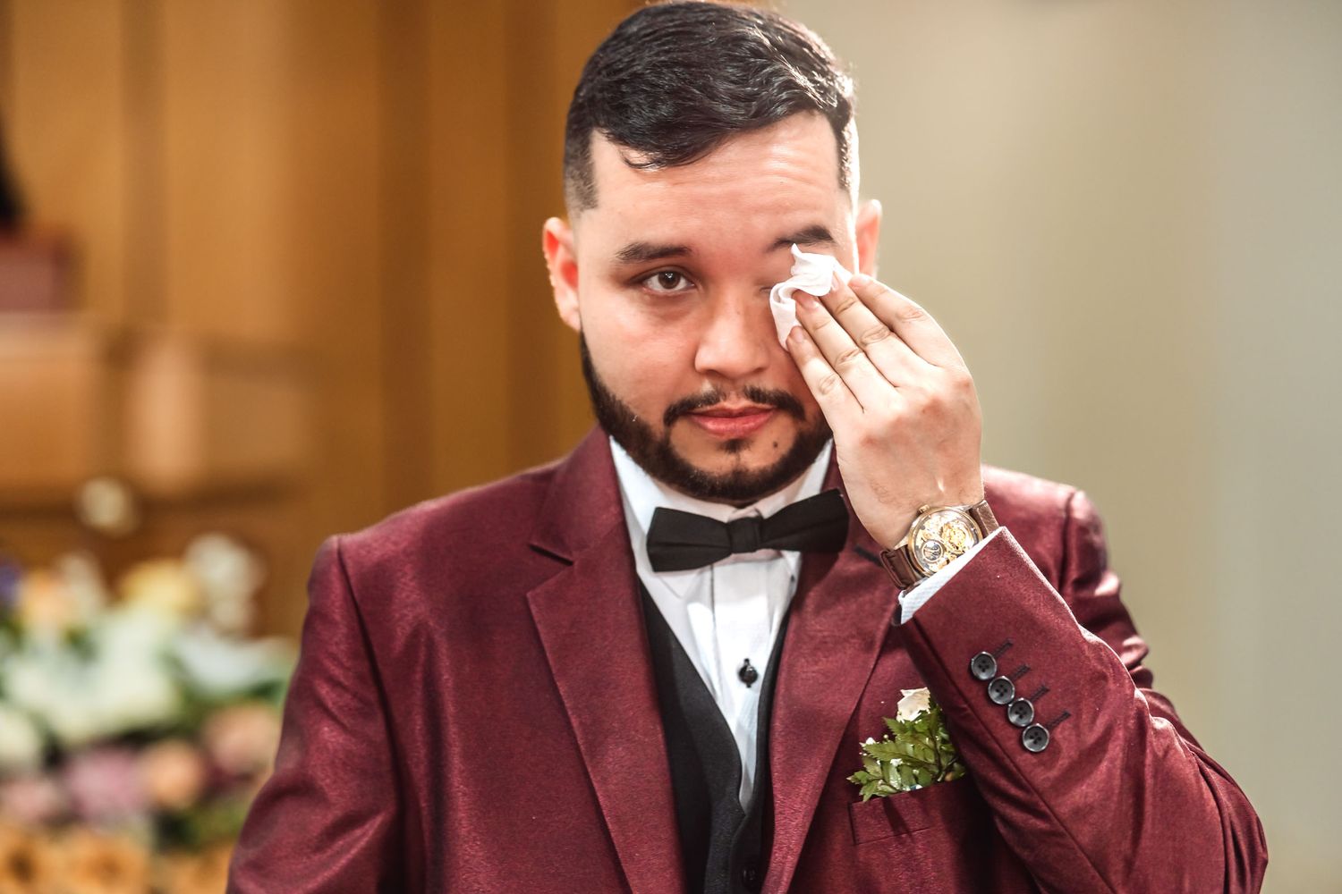 Novio con esmoquin borgoña y pajarita negra se seca lágrima con pañuelo; momento emotivo boda.