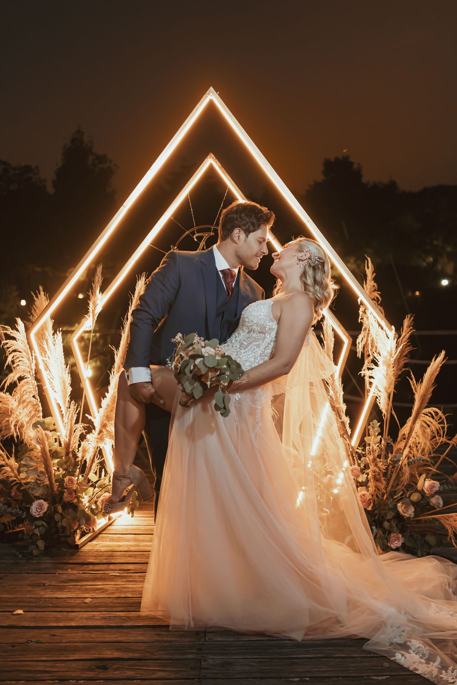 Novio levanta a novia (vestido rosa, ramo, velo) sonrientes bajo arco neón geométrico y pampas de noche. Boda.