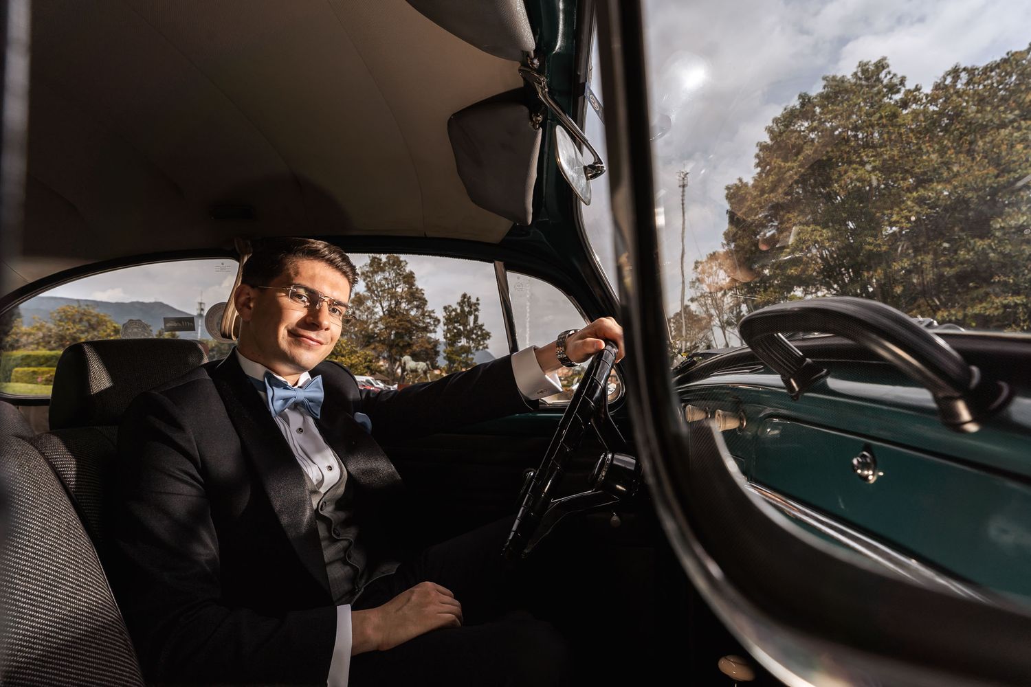 Novio elegante con esmoquin y pajarita azul sonríe al volante de coche clásico VW Escarabajo verde. Fotografía boda