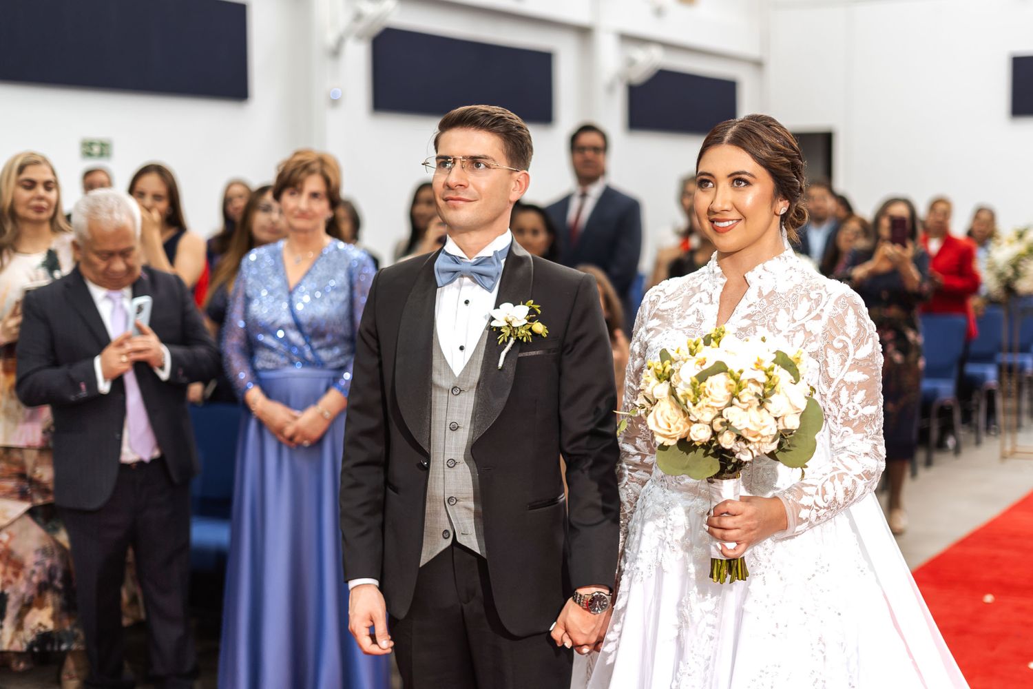 Novios sonríen de pie en pasillo rojo interior durante boda IDMJI, tomados de la mano, con invitados detrás.