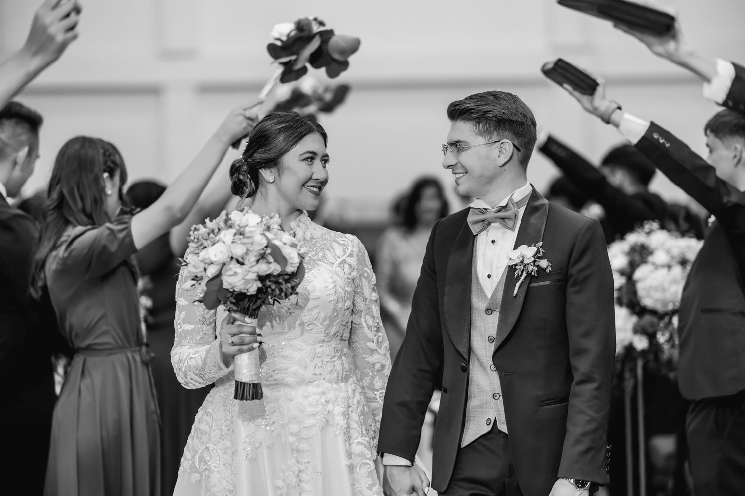 Novios caminan por pasillo en blanco y negro, mirándose y riendo. Invitados celebran su paso. Boda IDMJI.