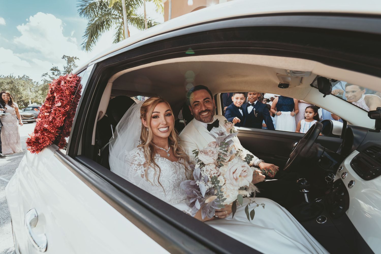 Novios sonrientes dentro de coche blanco; él camisa floral, ella vestido novia y velo. Flores rojas en ventana.