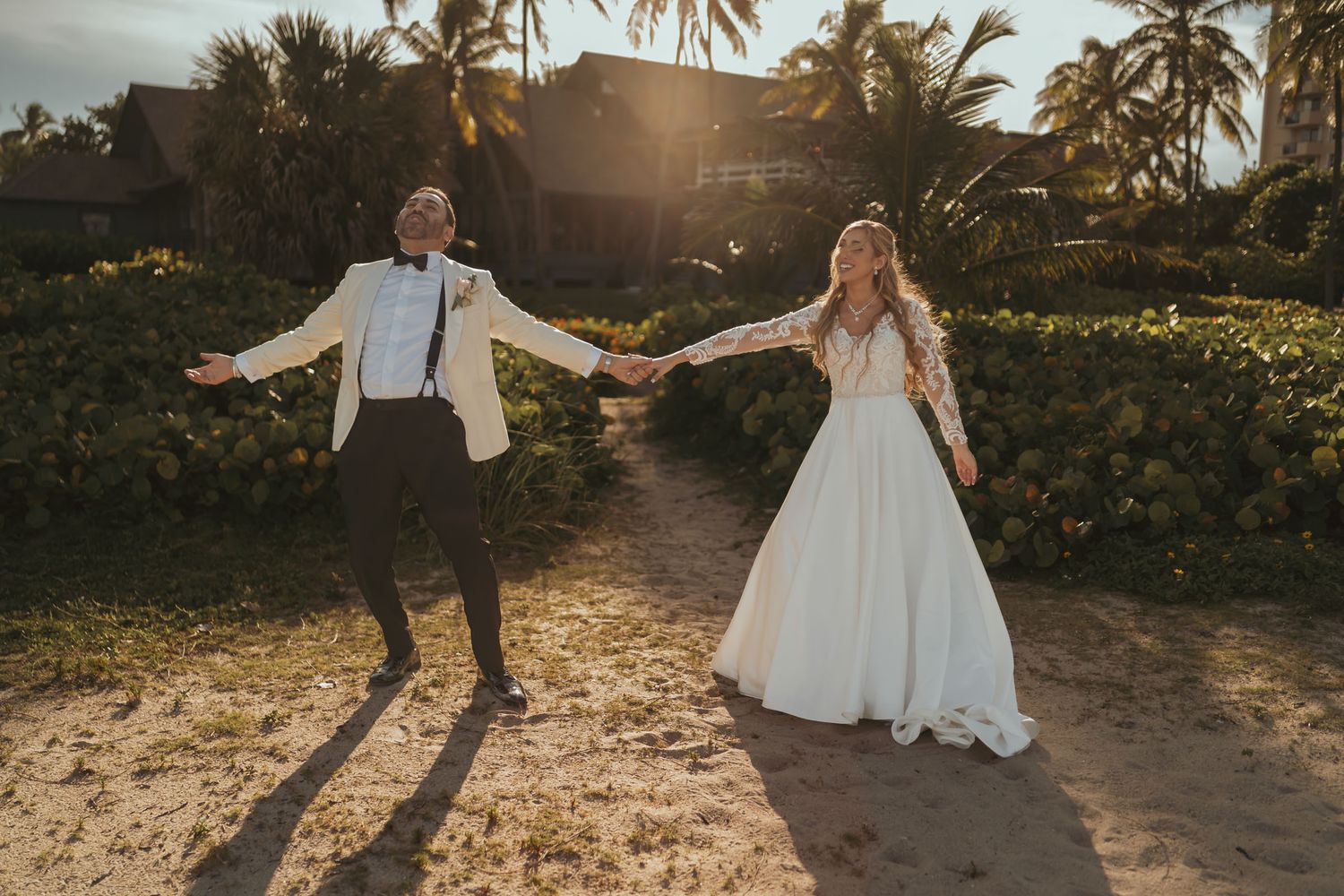 Novios felices tomados de la mano con brazos abiertos en camino de playa; sol brillante, palmeras. Boda destino.