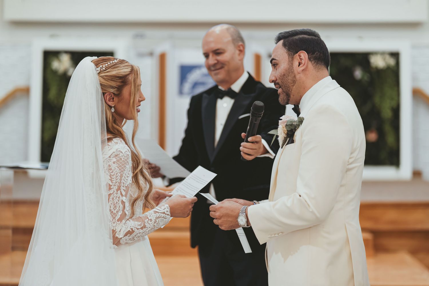 Novios y oficiante ríen juntos durante ceremonia de boda; novia sostiene los votos. Momento alegre.