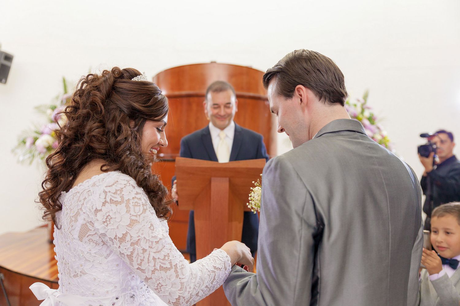 Novios intercambian anillos sonrientes durante ceremonia boda; oficiante sonríe al fondo en podio.