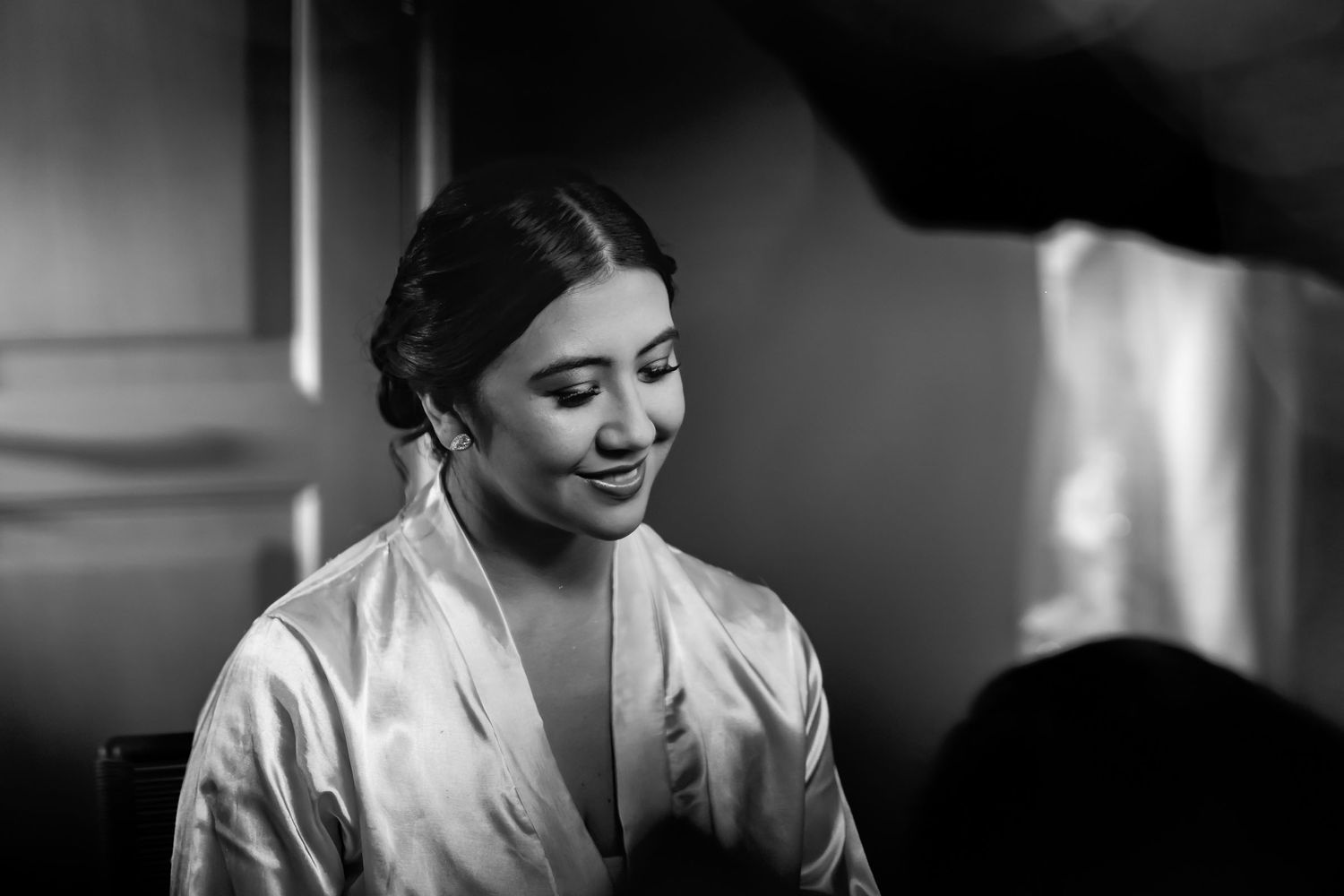 Foto blanco y negro: Novia con bata de satín sonríe suavemente mirando hacia abajo durante preparación de boda.