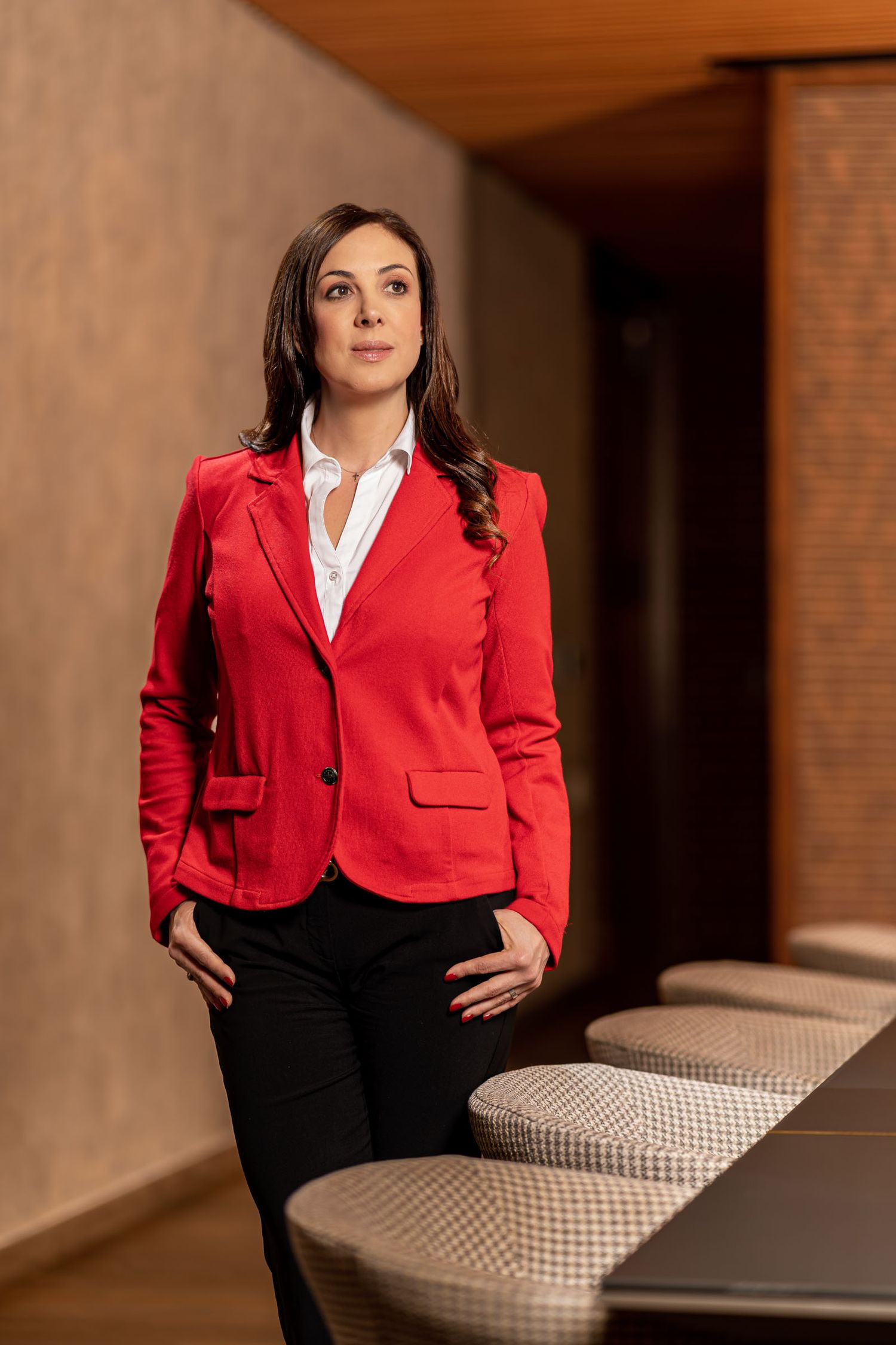 Mujer elegante con blazer rojo y camisa blanca posa de pie en interior moderno mirando hacia arriba. Sandra Vélez
