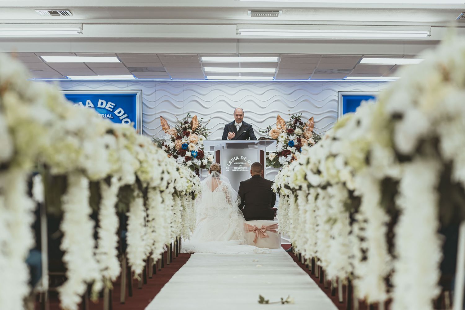 Vista por pasillo decorado hacia novios sentados y oficiante en podio durante ceremonia boda IDMJI.
