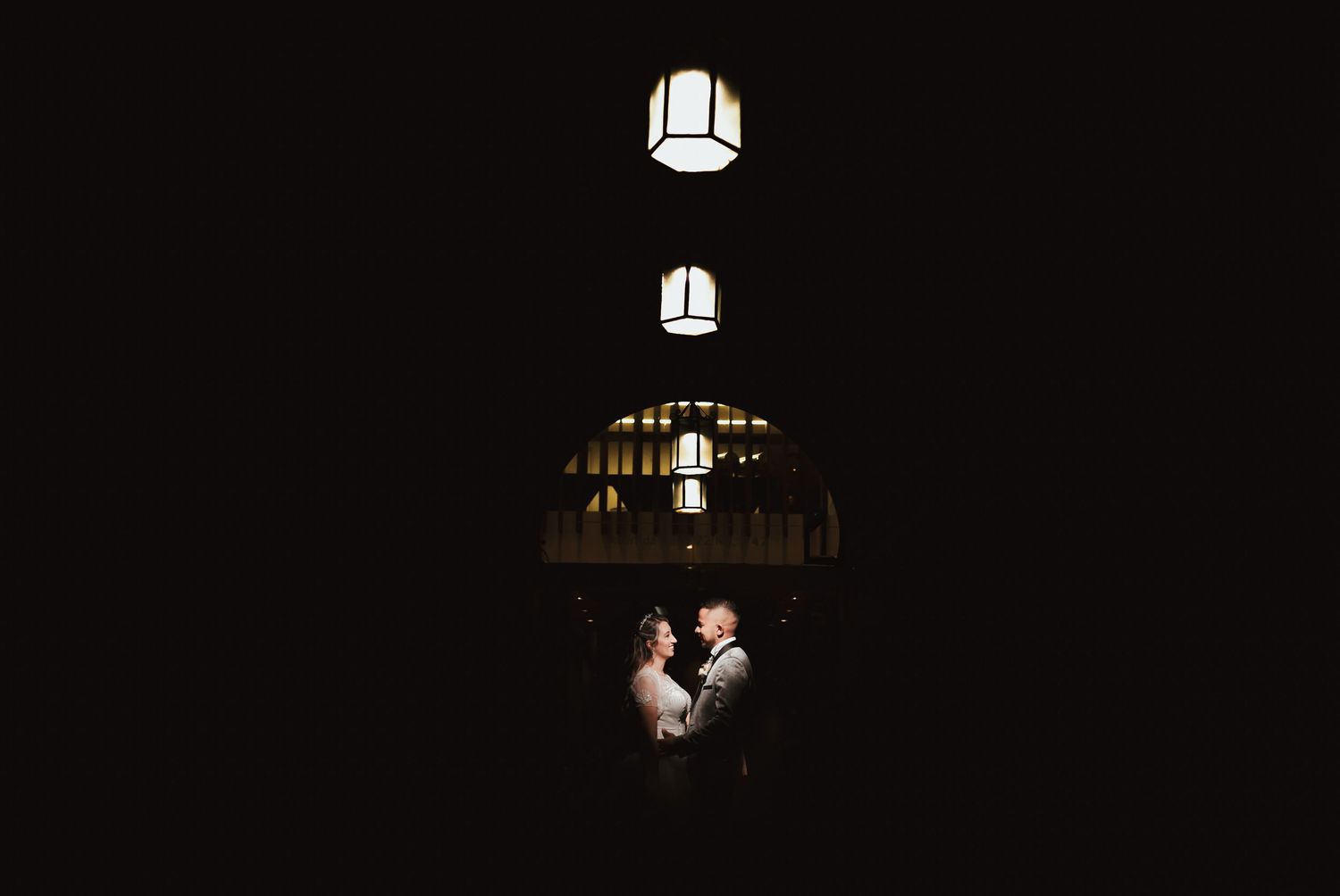 Pareja de novios se abraza en la oscuridad, iluminada por tres lámparas geométricas colgantes. Foto de boda íntima.