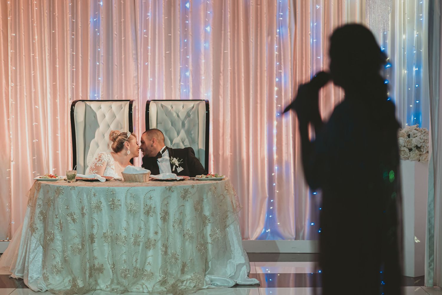 Silueta de cantante actúa mientras novios se besan en mesa principal; recepción boda, cortina luces.