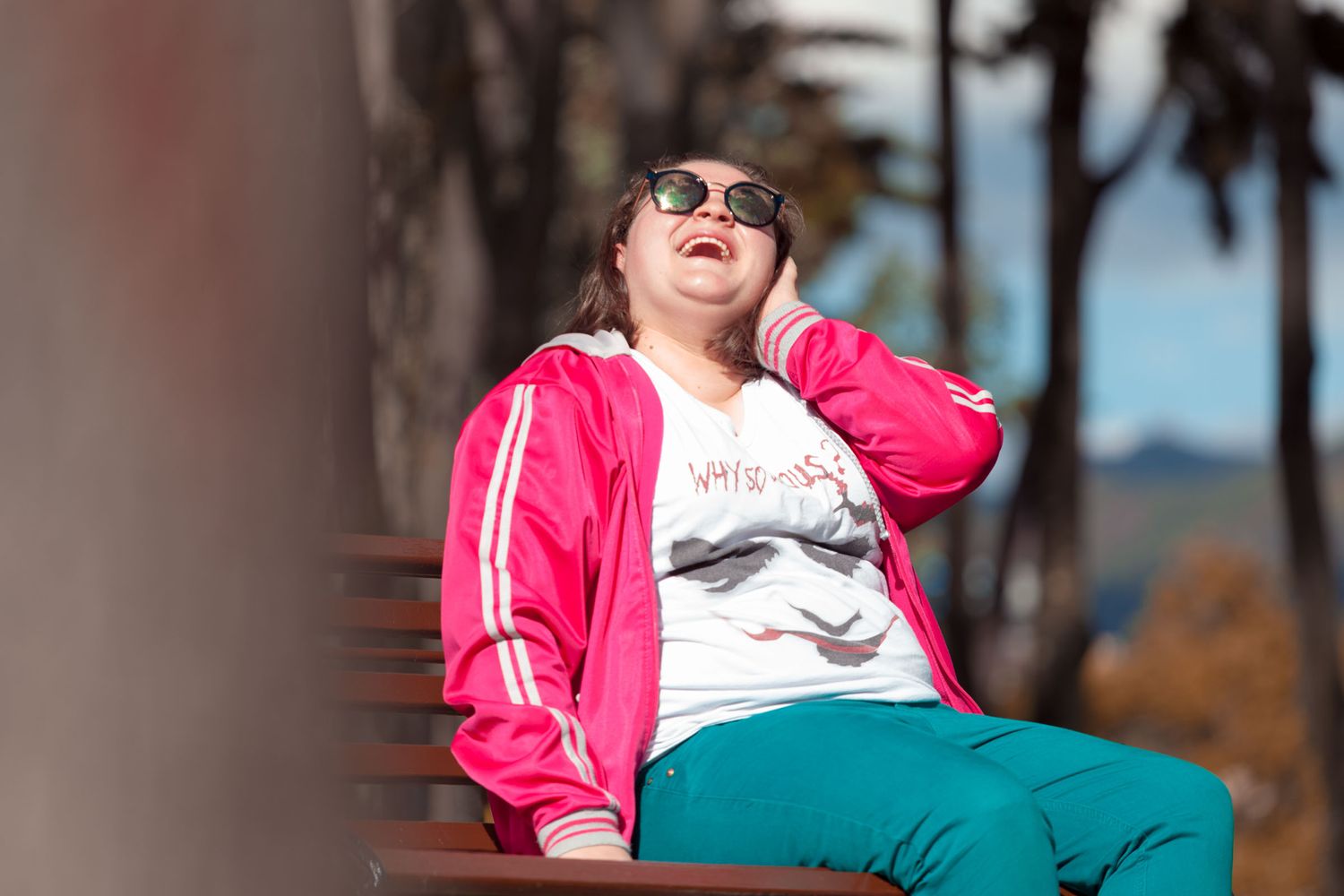 Persona joven con chaqueta fucsia y gafas de sol ríe a carcajadas sentada en banco exterior.
Sesión de 15
