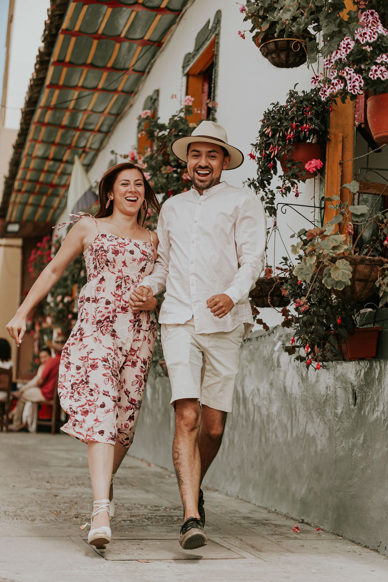 Pareja camina feliz por calle con flores en Salento. Ella viste vestido floral, él camisa y sombrero. Momento casual.
