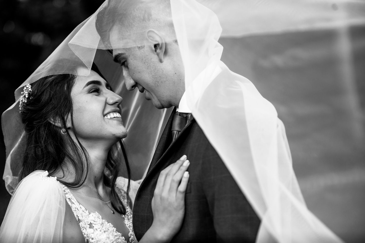 Foto B&W: Novia sonriente mira a novio bajo velo transparente; retrato íntimo boda.