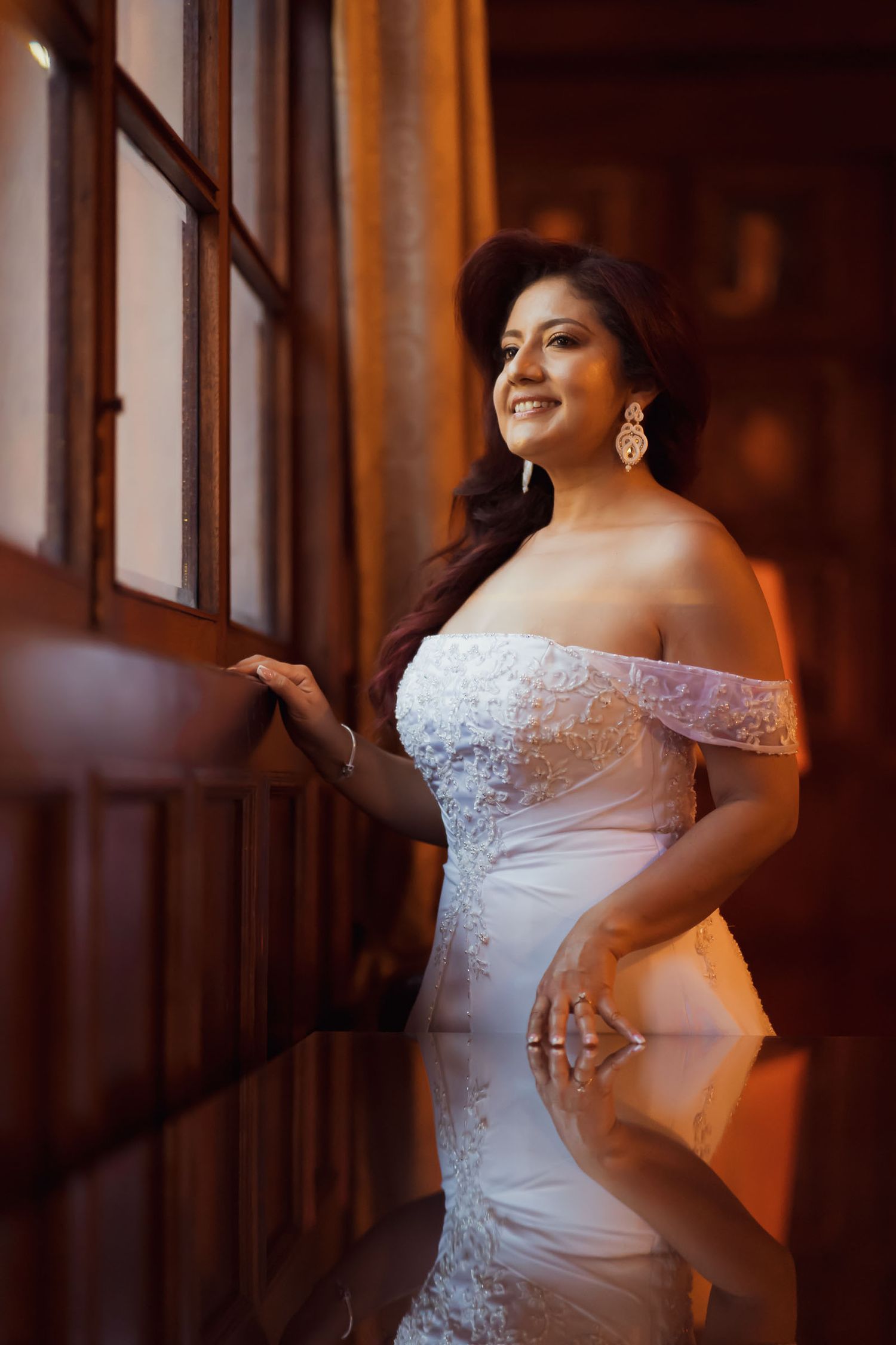 Novia con vestido blanco hombros descubiertos mira por ventana con luz cálida; retrato elegante boda.