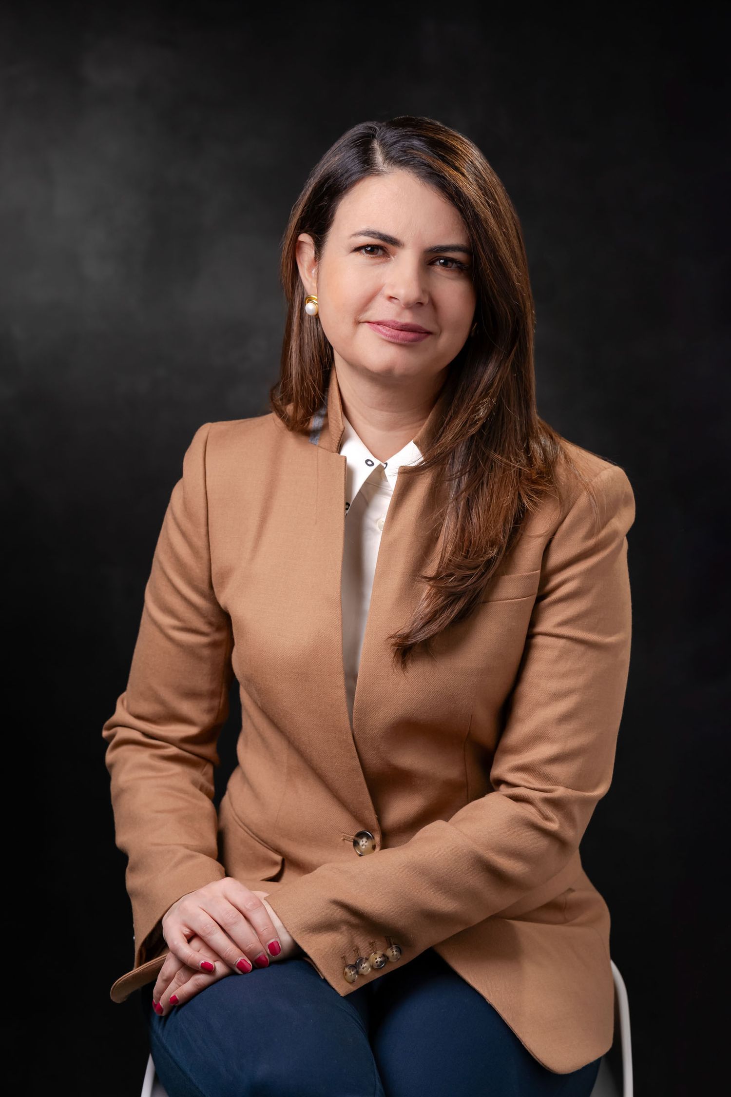 Retrato profesional mujer sentada; lleva blazer camel sobre camisa blanca, mira amable a cámara. Fondo oscuro.