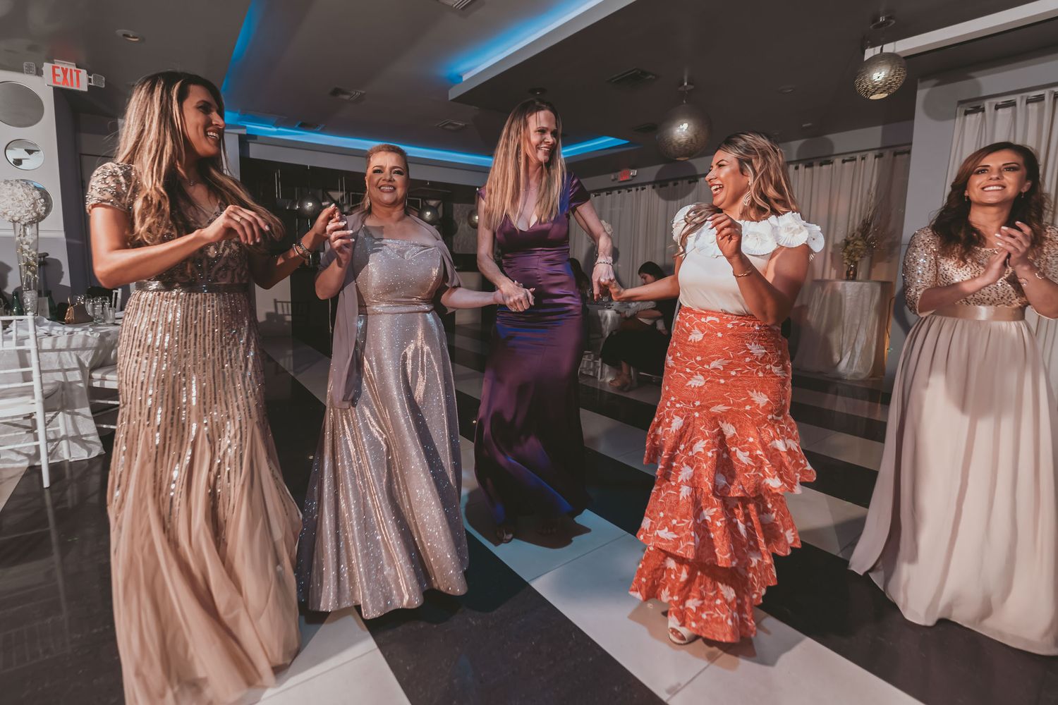 Grupo de mujeres invitadas con vestidos elegantes bailan juntas en pista a cuadros; fiesta de boda animada.