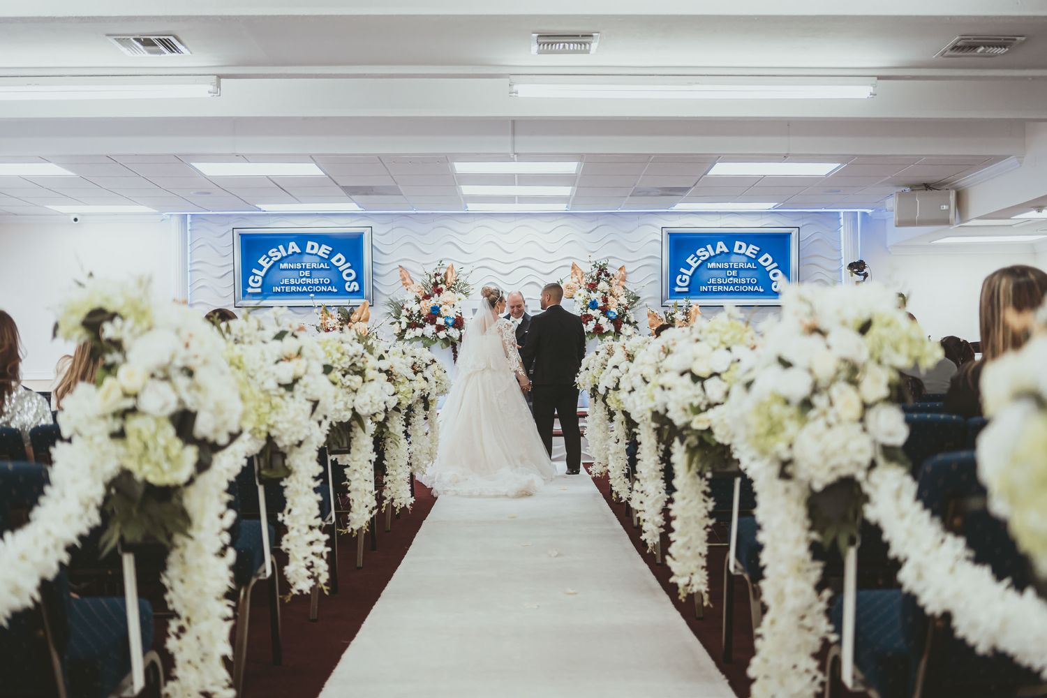Vista desde entrada de iglesia hacia novios en altar; largo pasillo blanco decorado con flores blancas. Boda IDMJI.