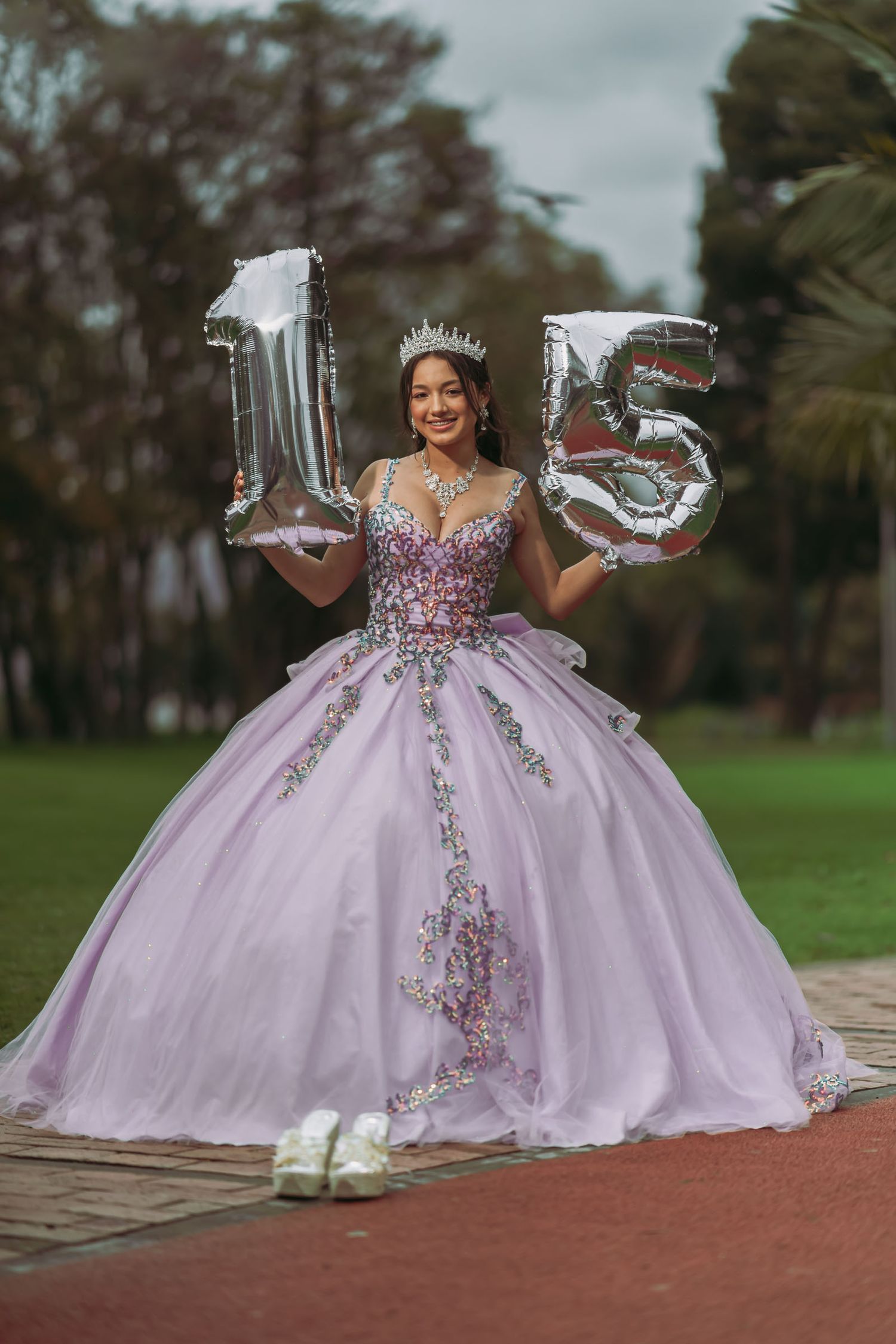 Quinceañera con vestido princesa lavanda y tiara sostiene globos plateados gigantes 