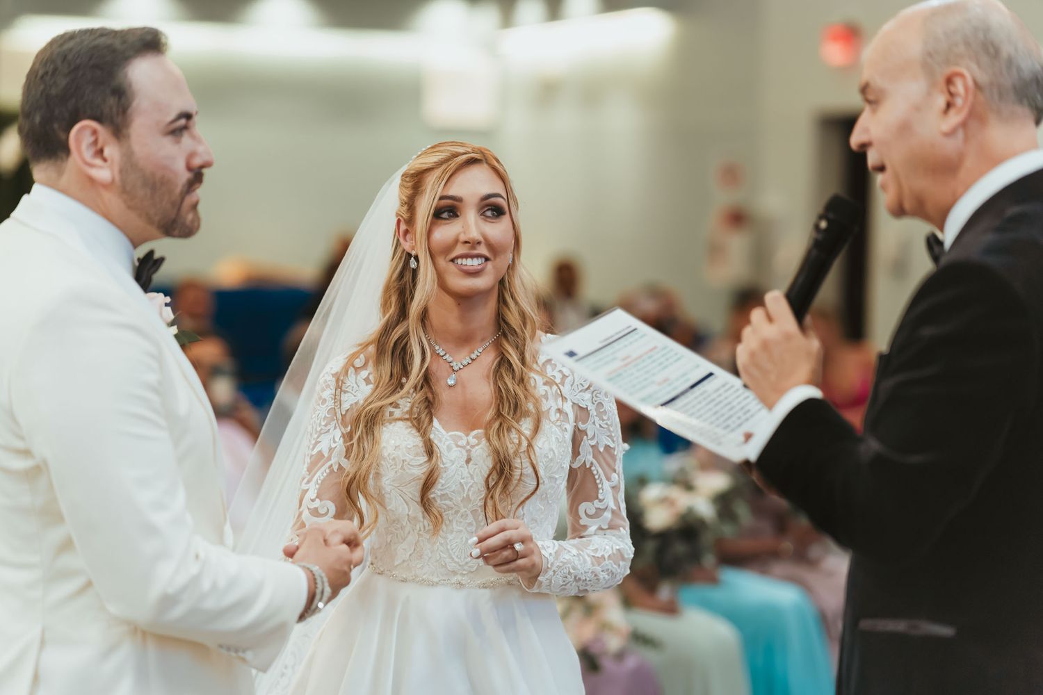 Novios tomados de la mano miran a oficiante que habla con micrófono durante ceremonia boda; votos.