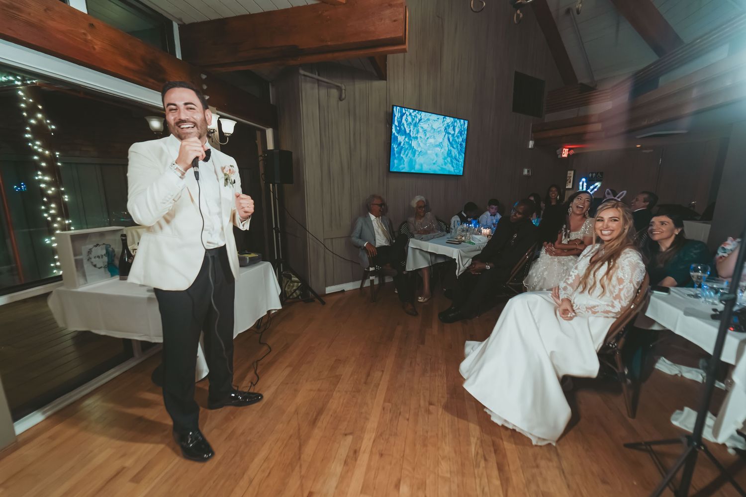 Novio con chaqueta blanca da discurso o canta con micrófono en recepción; novia sentada mira sonriente.