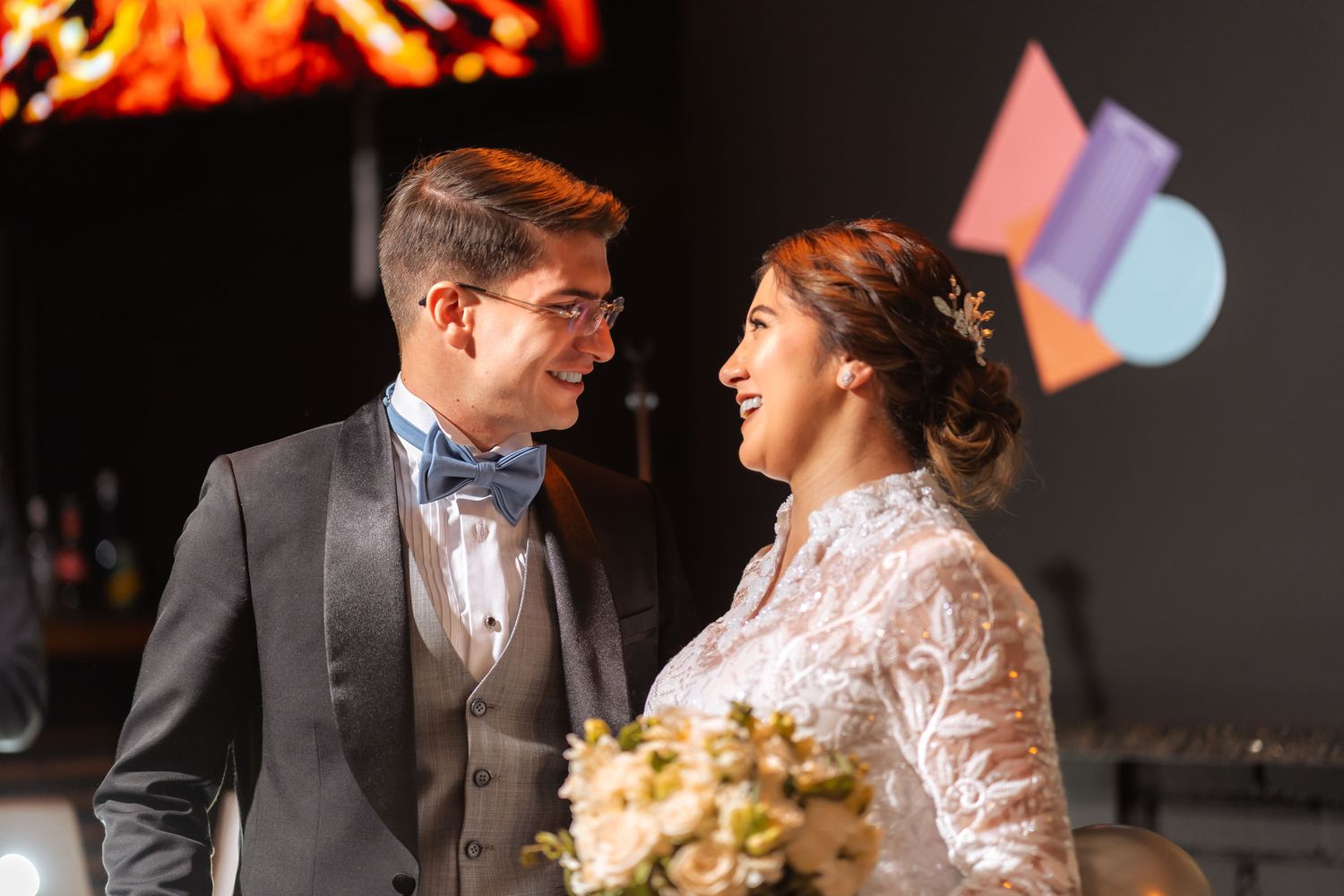 Novios se miran y sonríen/ríen felices en escenario con decoración geométrica colorida. Foto de boda.