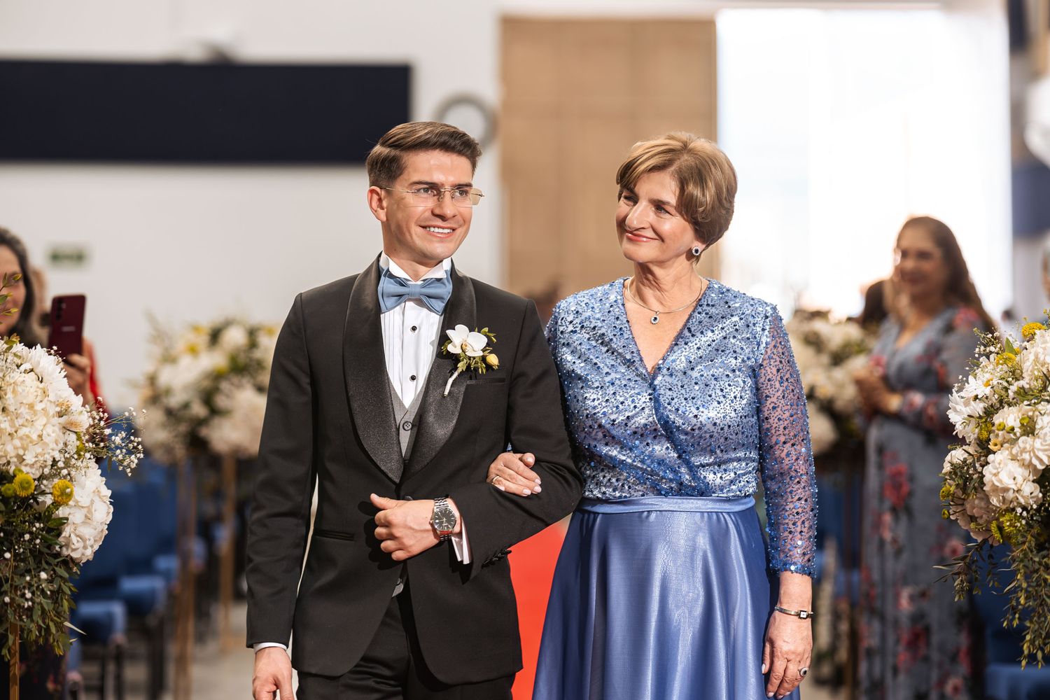 Novio sonriente con esmoquin y pajarita azul mira a su madre mientras caminan juntos por el pasillo en la boda.