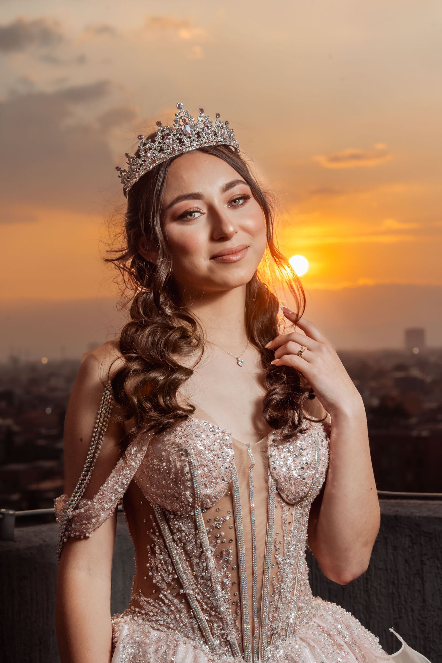 Primer plano: Quinceañera con vestido corsé rosa y tiara posa suavemente frente al sol poniente sobre la ciudad.