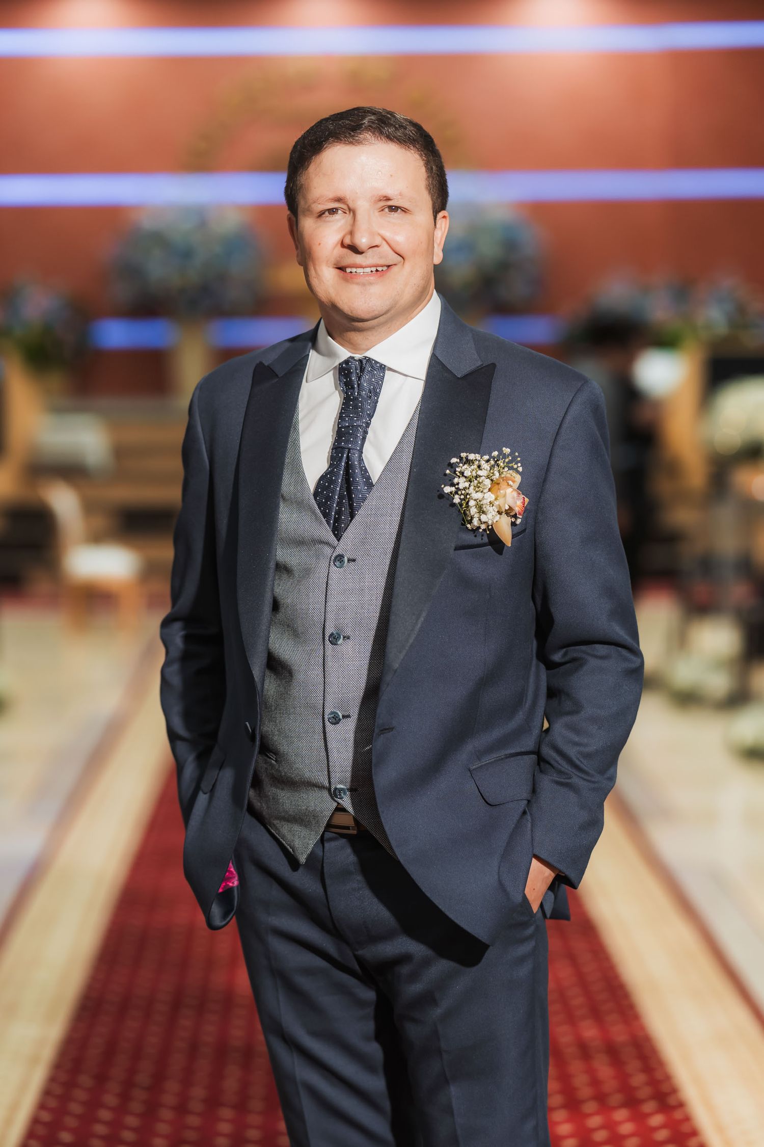 Retrato novio sonriente con traje azul tres piezas y boutonnière posa en pasillo rojo de iglesia. Fotografía boda.