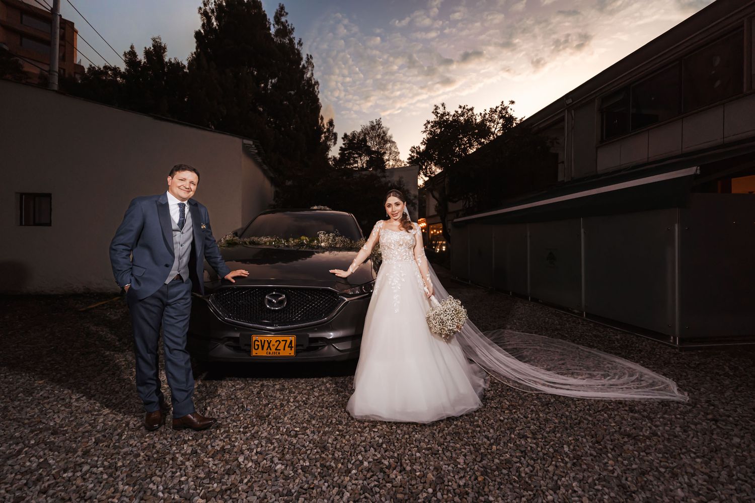 Novia y novio posan sonrientes frente a coche oscuro decorado para boda al atardecer; velo largo, ramo.