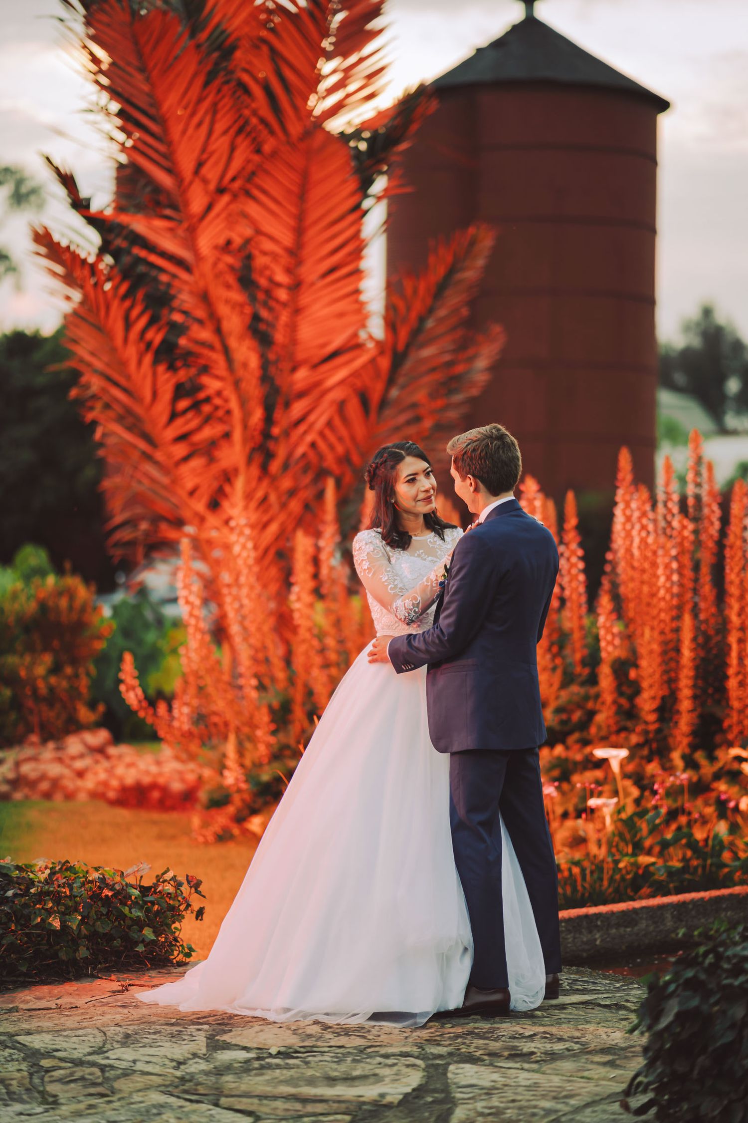 Novios abrazados en un jardín con una palmera iluminada de rojo en hacienda en Cajicá
