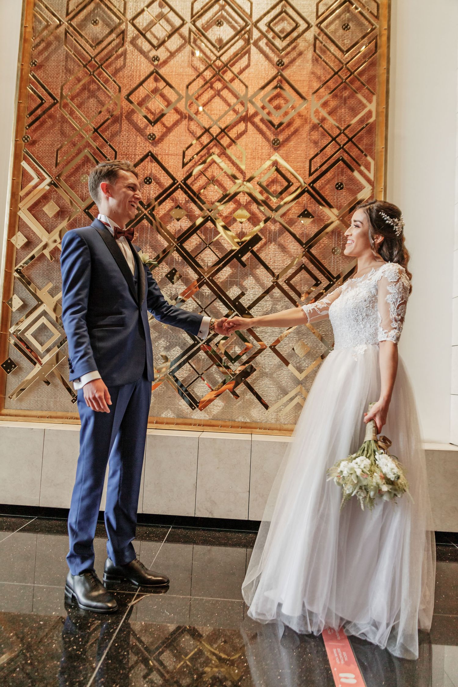 Novios se giran para verse, sonriendo; 'first look' boda frente a panel decorativo dorado. Novia ramo.