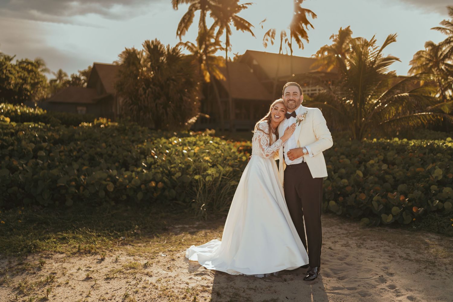 Novios se abrazan sonrientes en playa tropical al atardecer; palmeras y cabañas fondo. Boda romántica.
