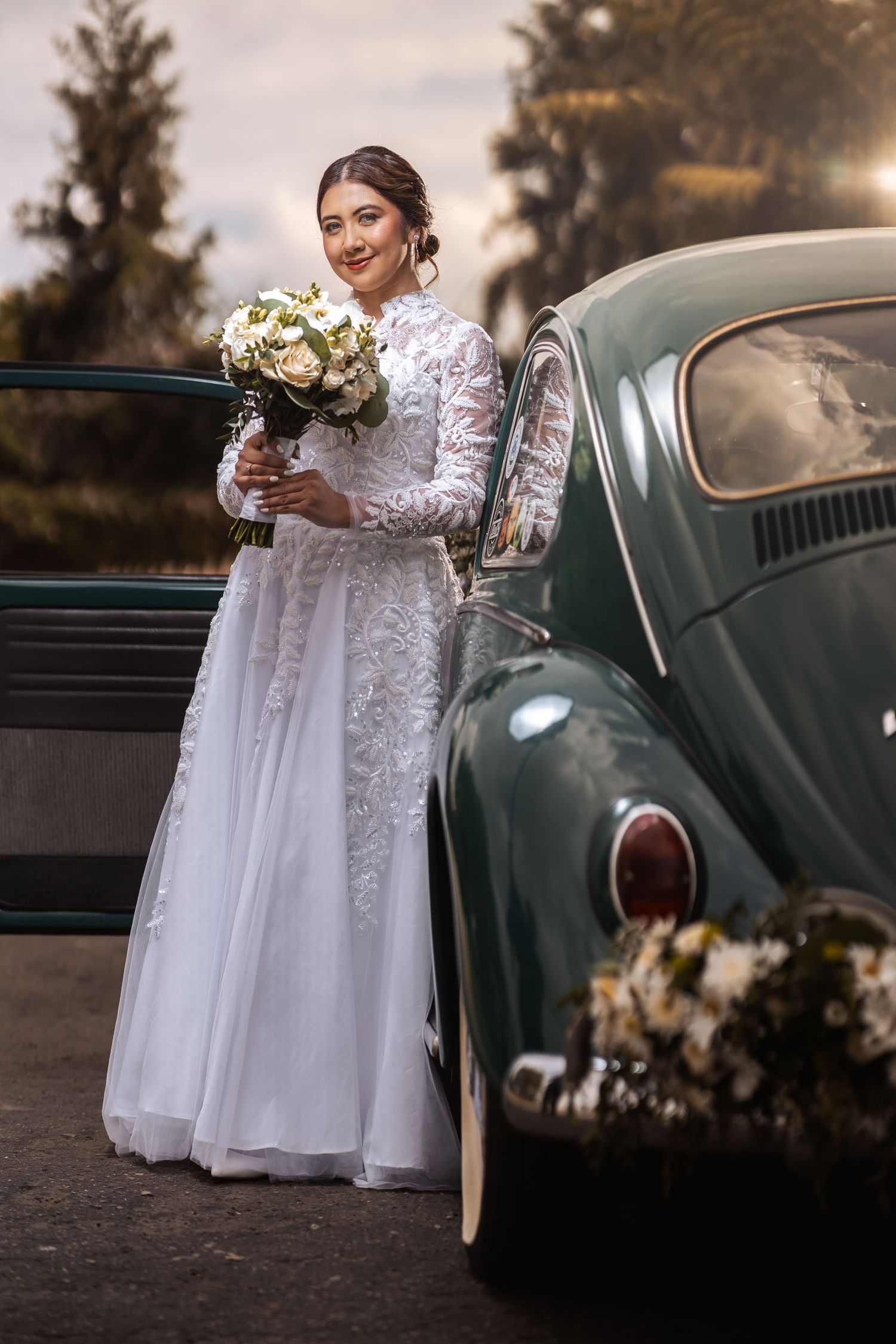 Novia posa elegante de cuerpo entero con vestido encaje blanco y ramo junto a coche clásico VW Escarabajo verde. Boda.