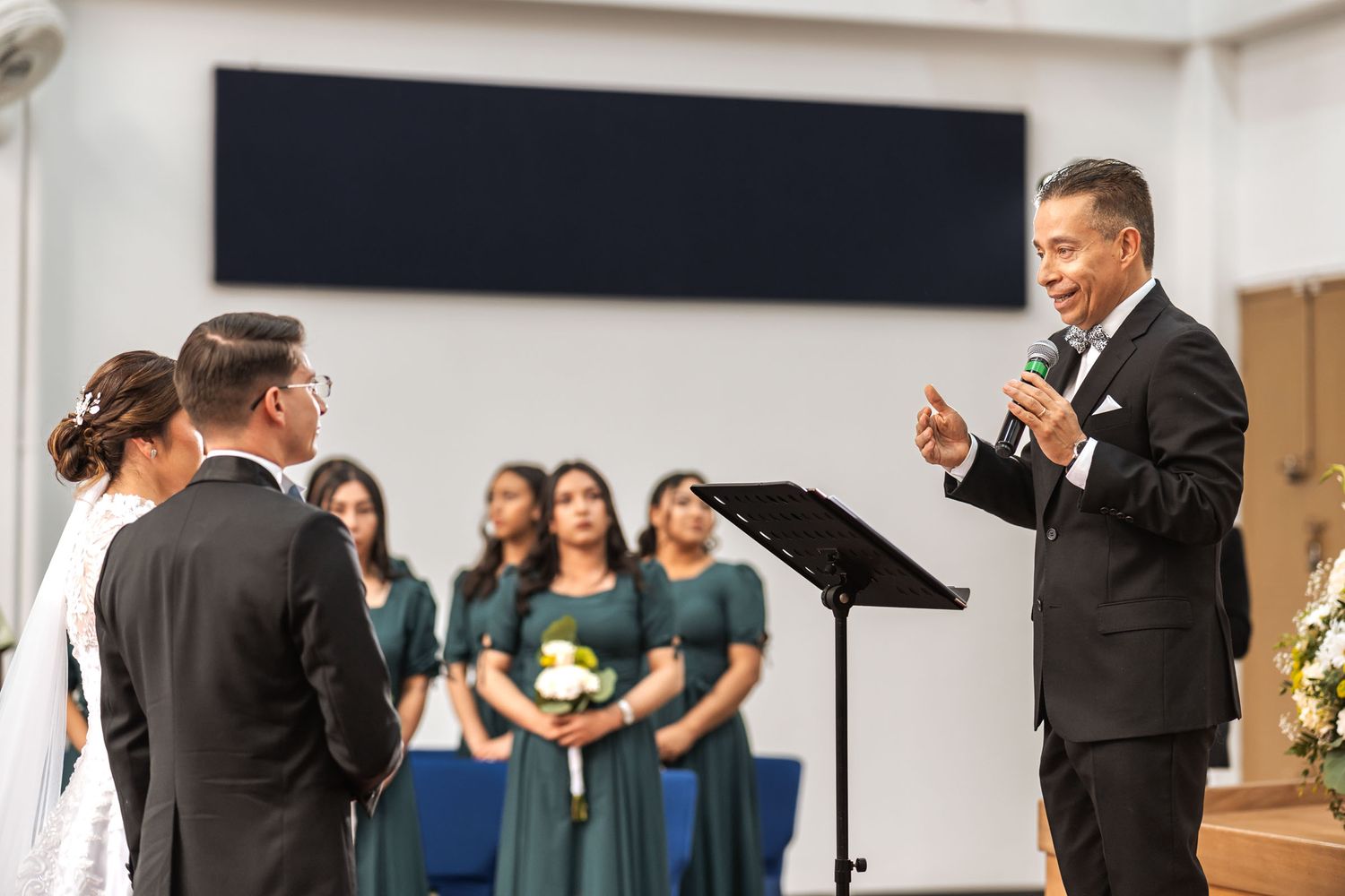 Novios sonríen abrazados en interior durante su boda frente al pastor que ministra la celebración. Comparten un momento feliz. Boda IDMJI