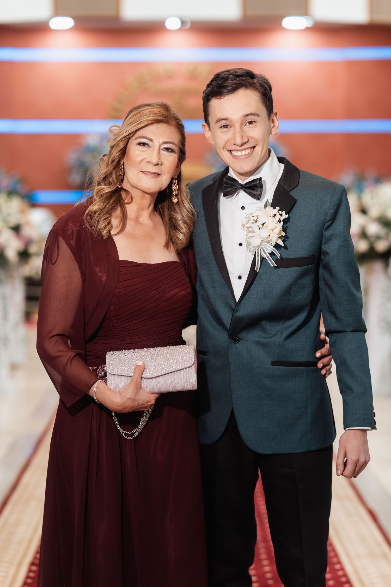 Novio sonriente con traje verde azulado junto a su madre en vestido borgoña.