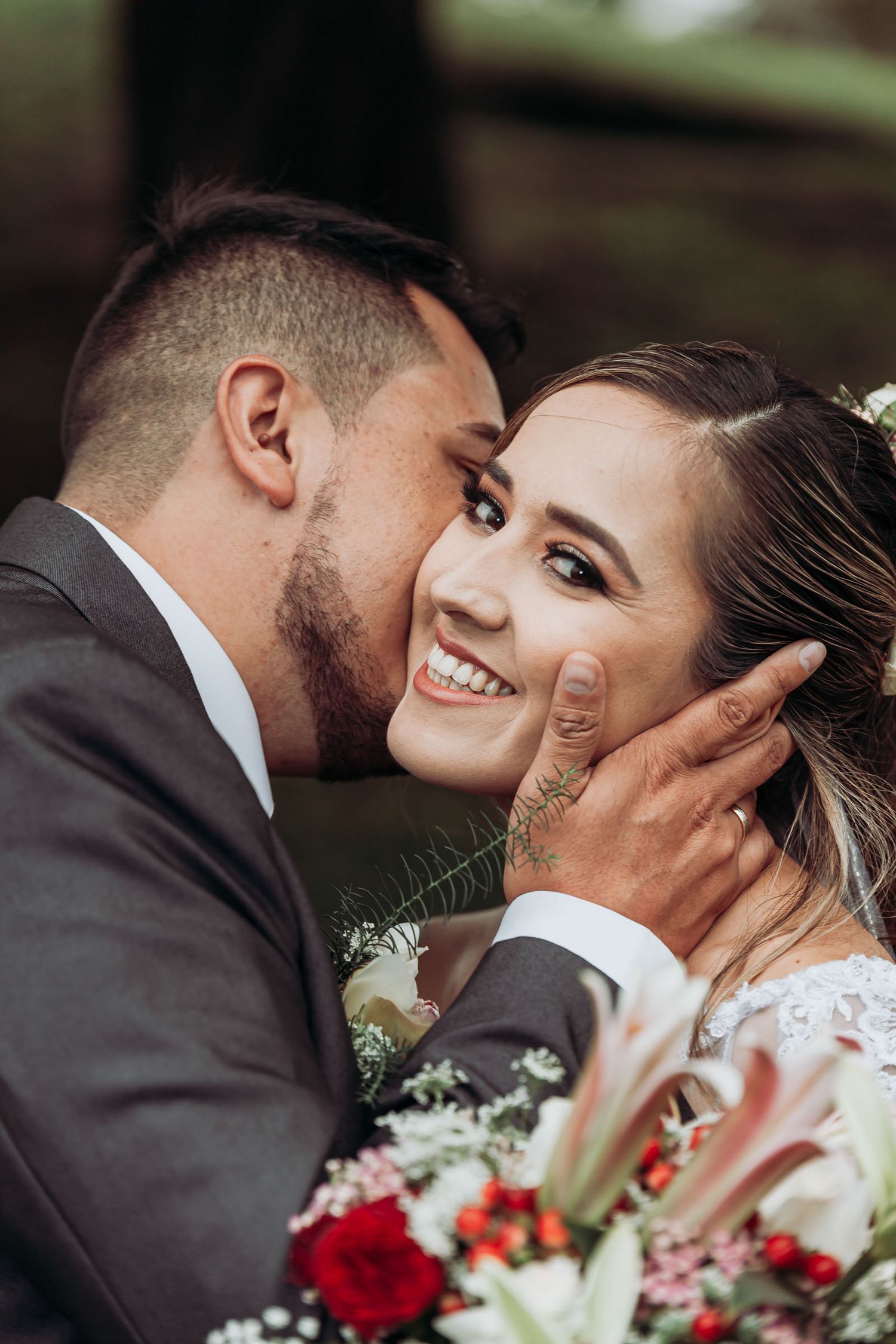 Novio besando a la novia en la mejilla y la novia sonriente con un ramo de flores en la mano