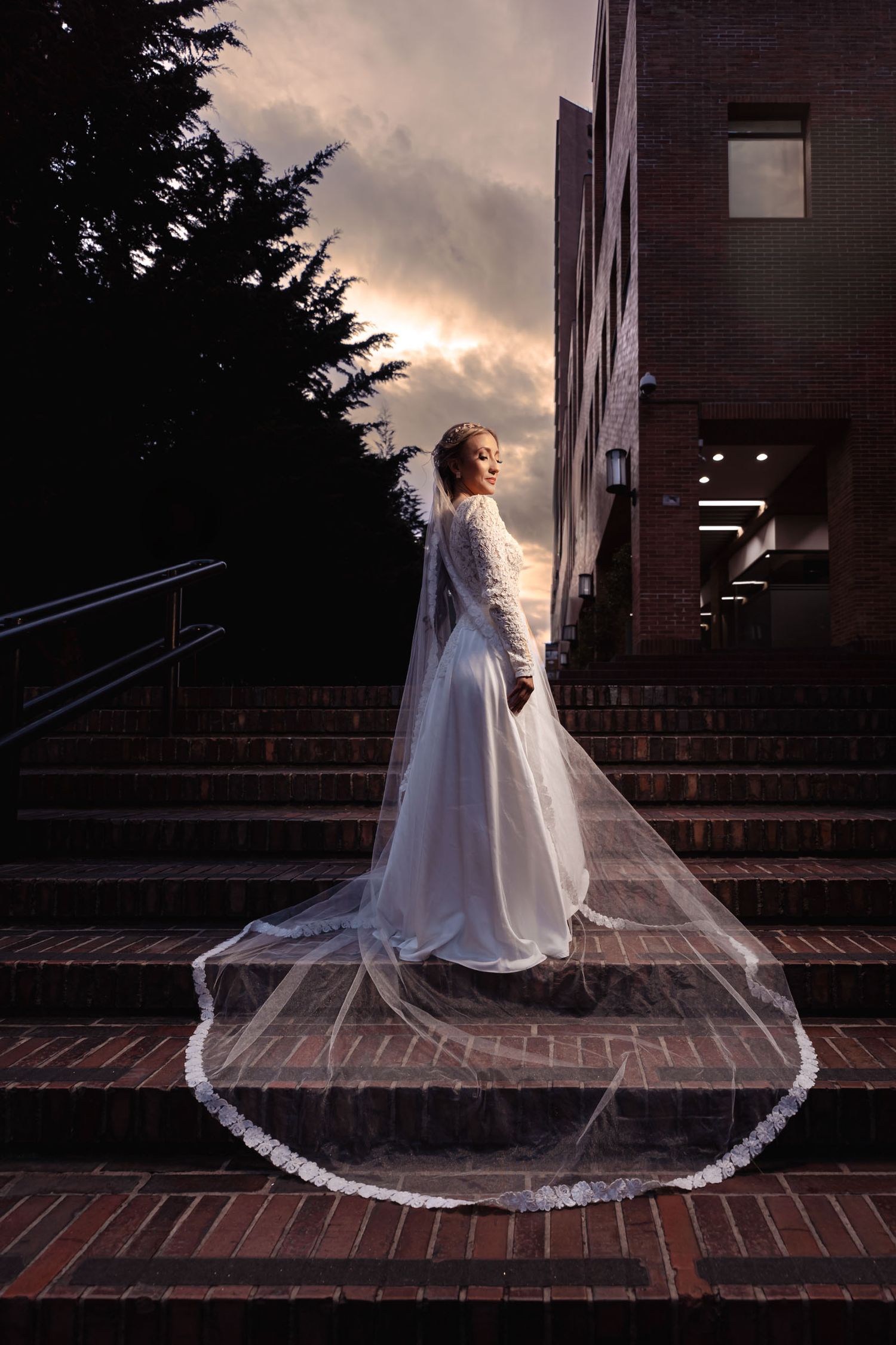 Novia elegante de espaldas en escaleras de ladrillo al atardecer. Viste vestido blanco y largo velo con detalles. Cielo dramático en ciudad