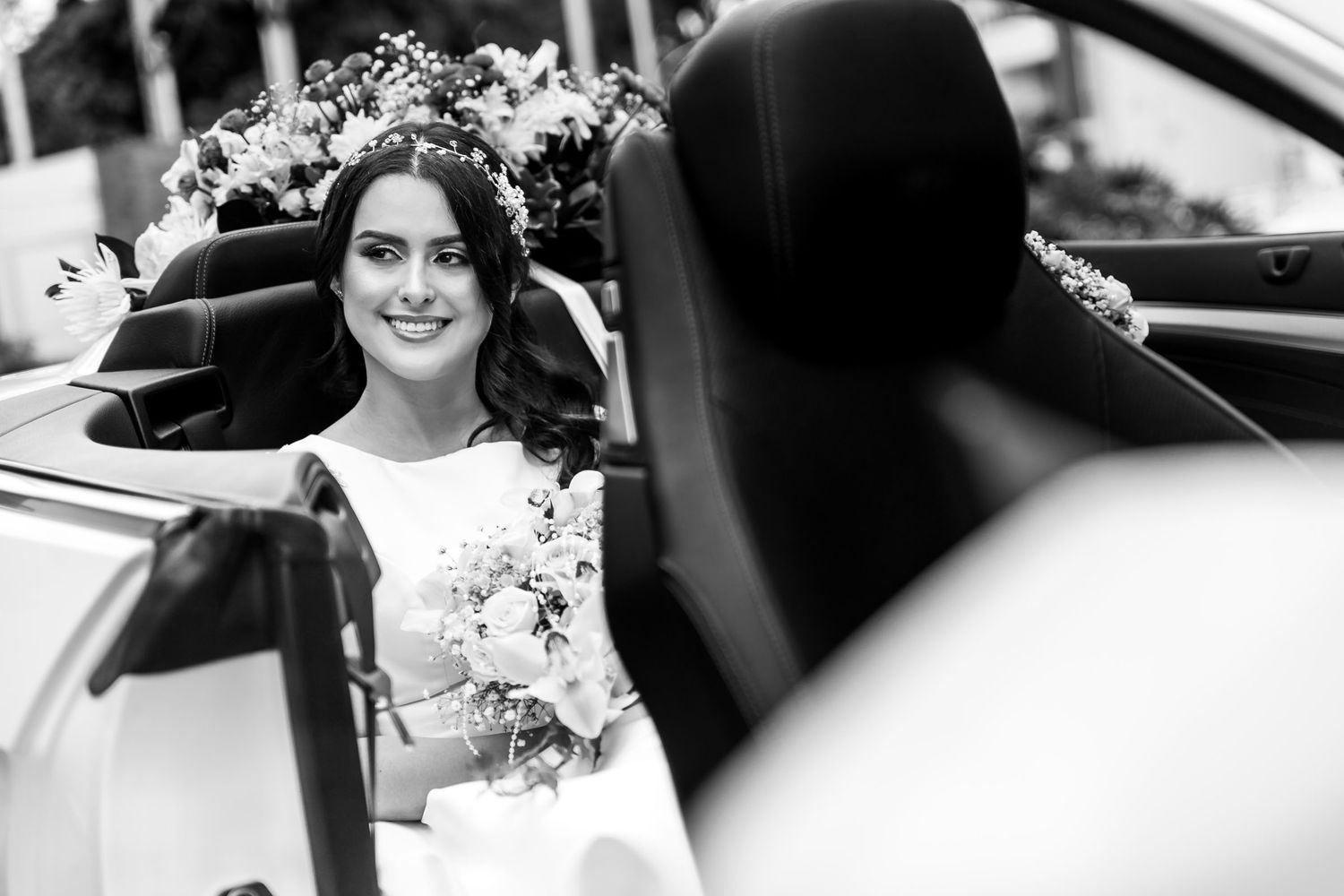 Foto B&W: Novia sonriente con ramo sentada en coche convertible clásico; retrato boda elegante.