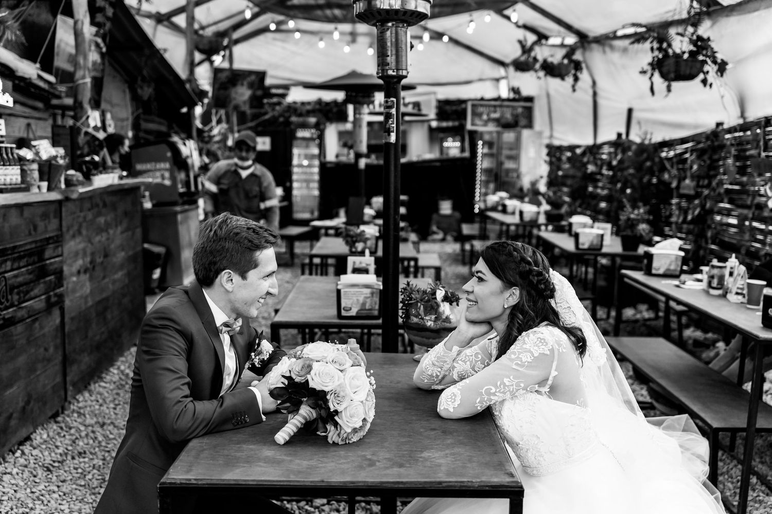 Foto B&W: Novios se miran sentados en mesa de restaurante; ramo sobre mesa. Boda urbana