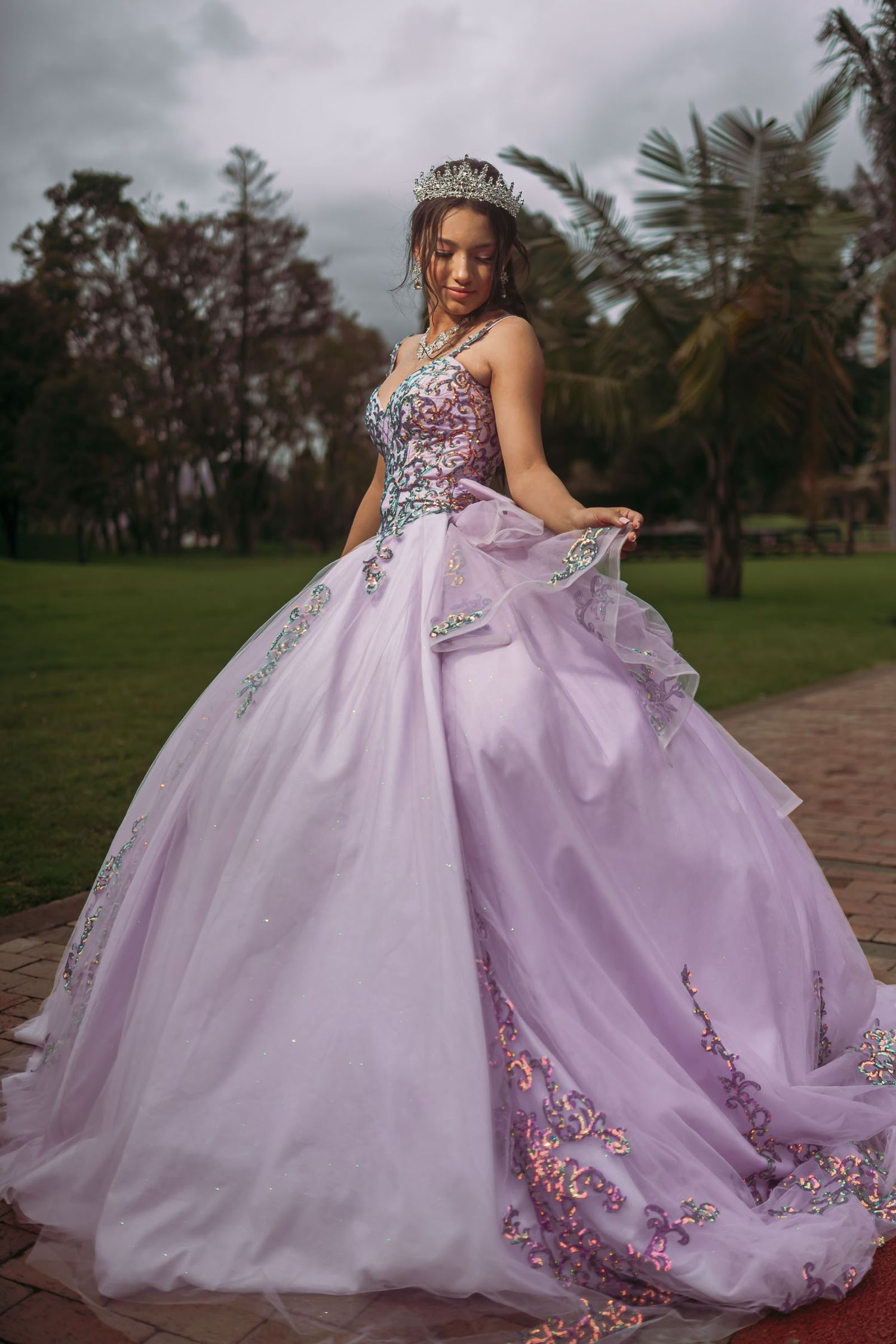 Quinceañera posa con vestido princesa lavanda (bordado flores) y tiara en parque; sesión fotos exterior.
