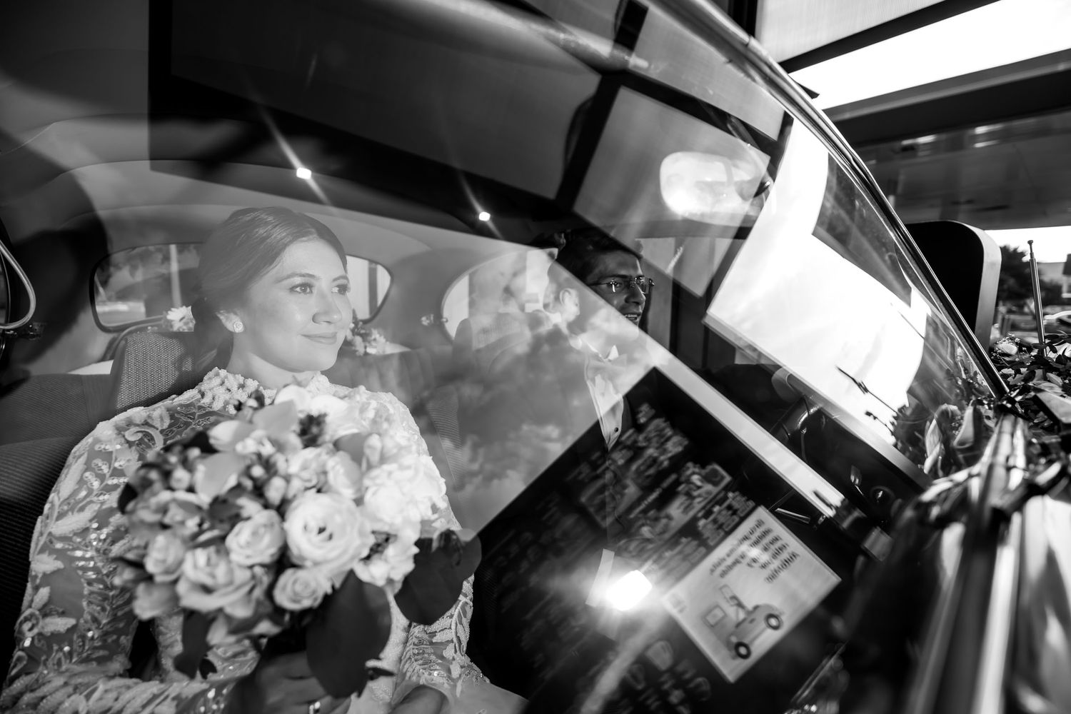 Novia sonríe sentada en coche, sosteniendo su ramo floral, en foto de boda blanco y negro. Momento íntimo.
