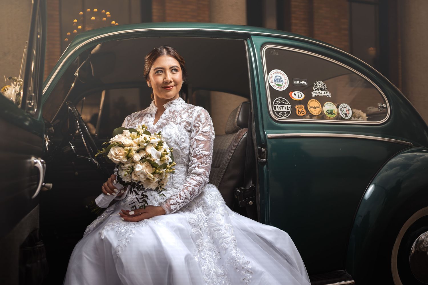 Novia sonriente con vestido de encaje y ramo sentada en la puerta abierta de un coche clásico VW Escarabajo verde. Boda.