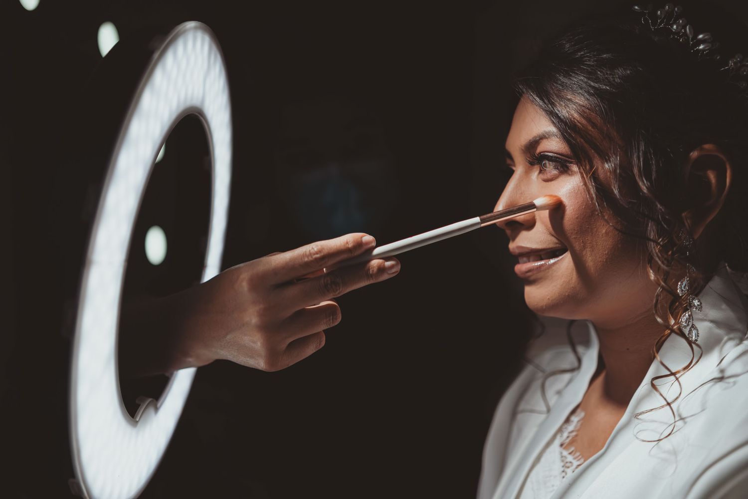 Primer plano: Mano aplicando maquillaje con brocha en rostro de mujer sonriente, iluminada por aro de luz. Preparación y alistamiento boda.