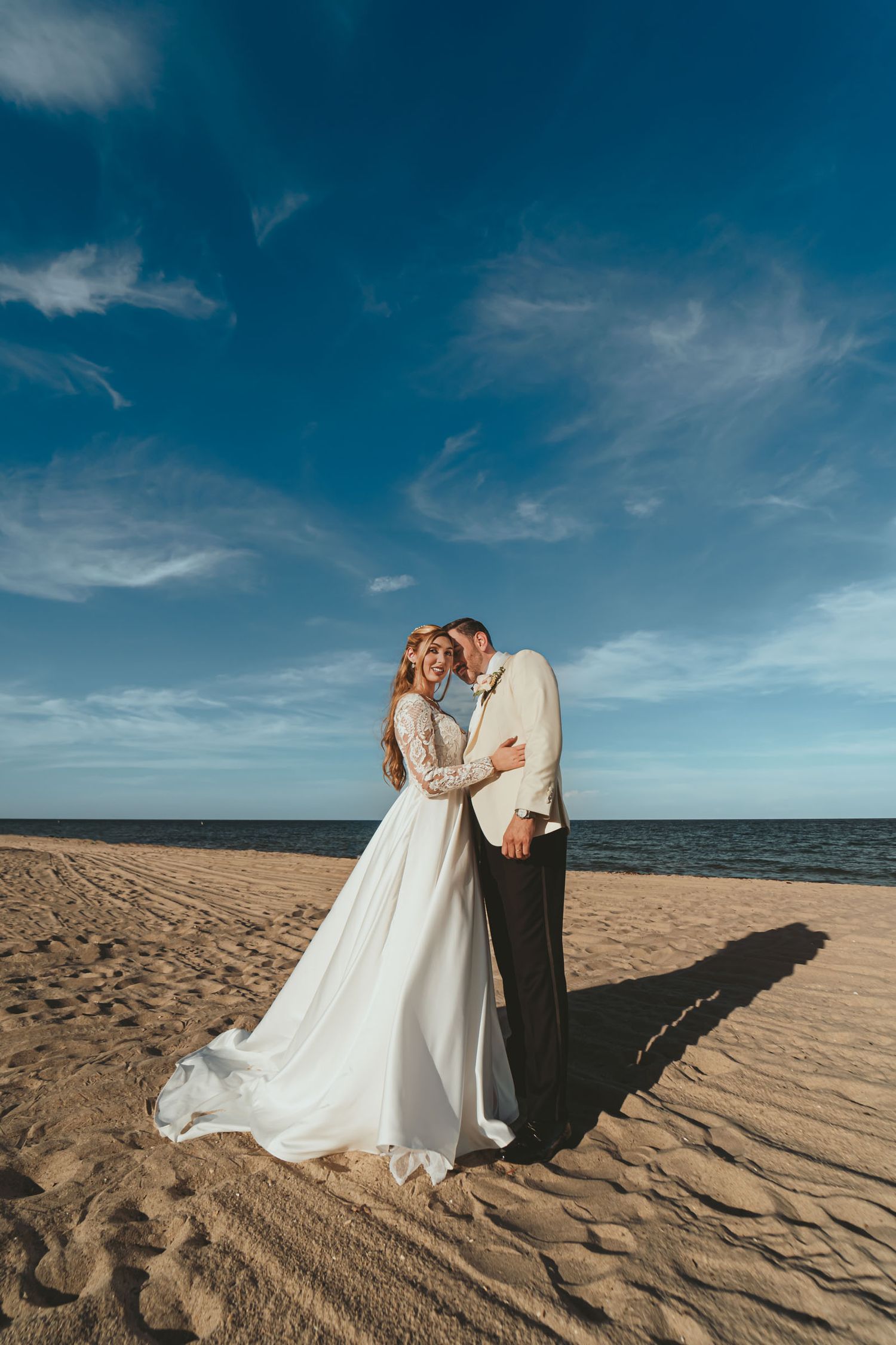 Novios se besan en playa de arena bajo cielo azul; él chaqueta clara, ella vestido blanco. Boda destino Miami.