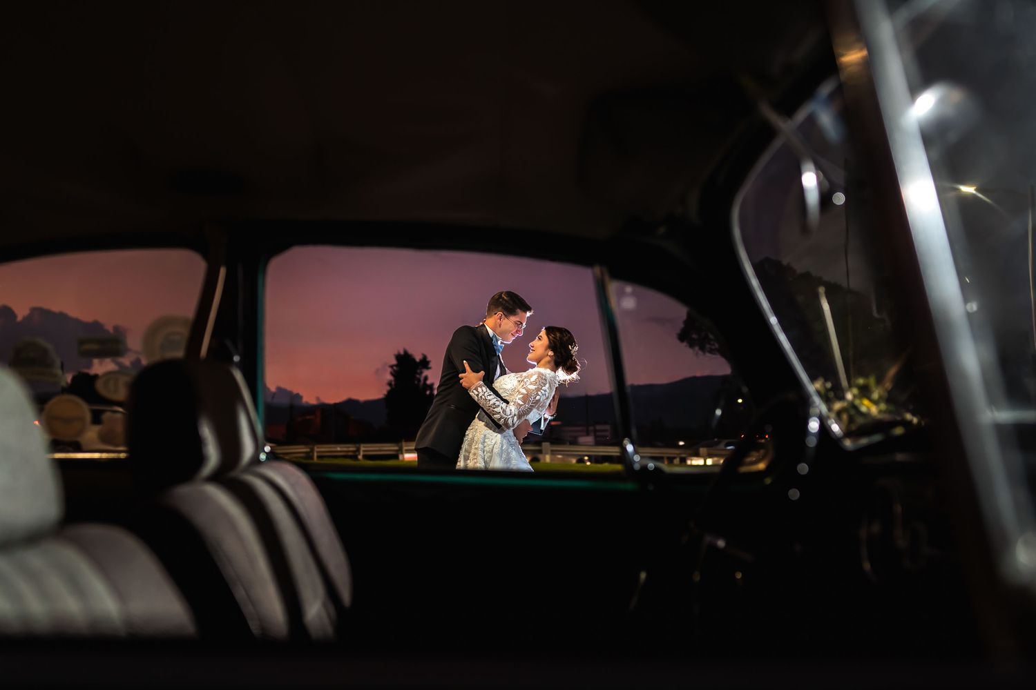 Novios se abrazan románticamente en exterior al atardecer, vistos a través de la ventana de un coche. Foto de boda.