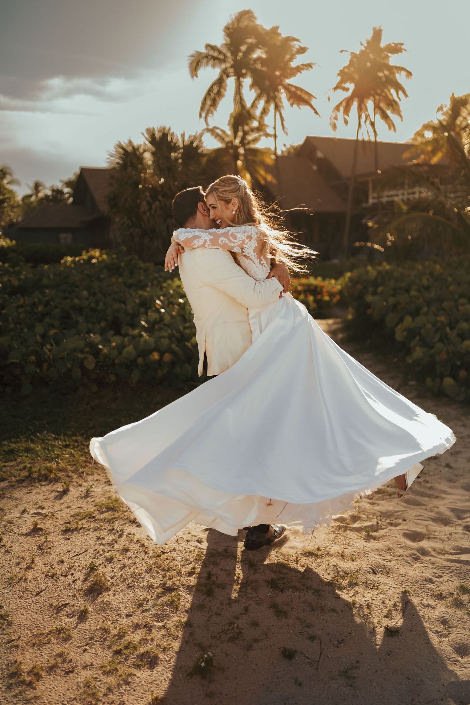 Novio con chaqueta clara levanta a novia (vestido blanco) en playa tropical; ella ríe. Atardecer, palmeras.