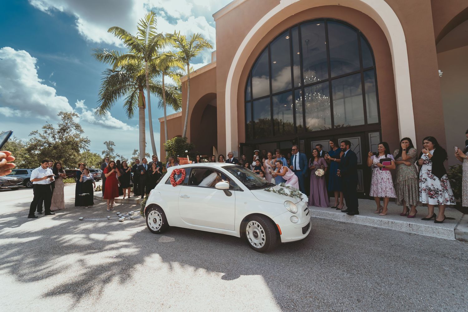 Pequeño coche blanco de boda llega a edificio con arco; invitados esperan bajo palmeras. Día soleado.