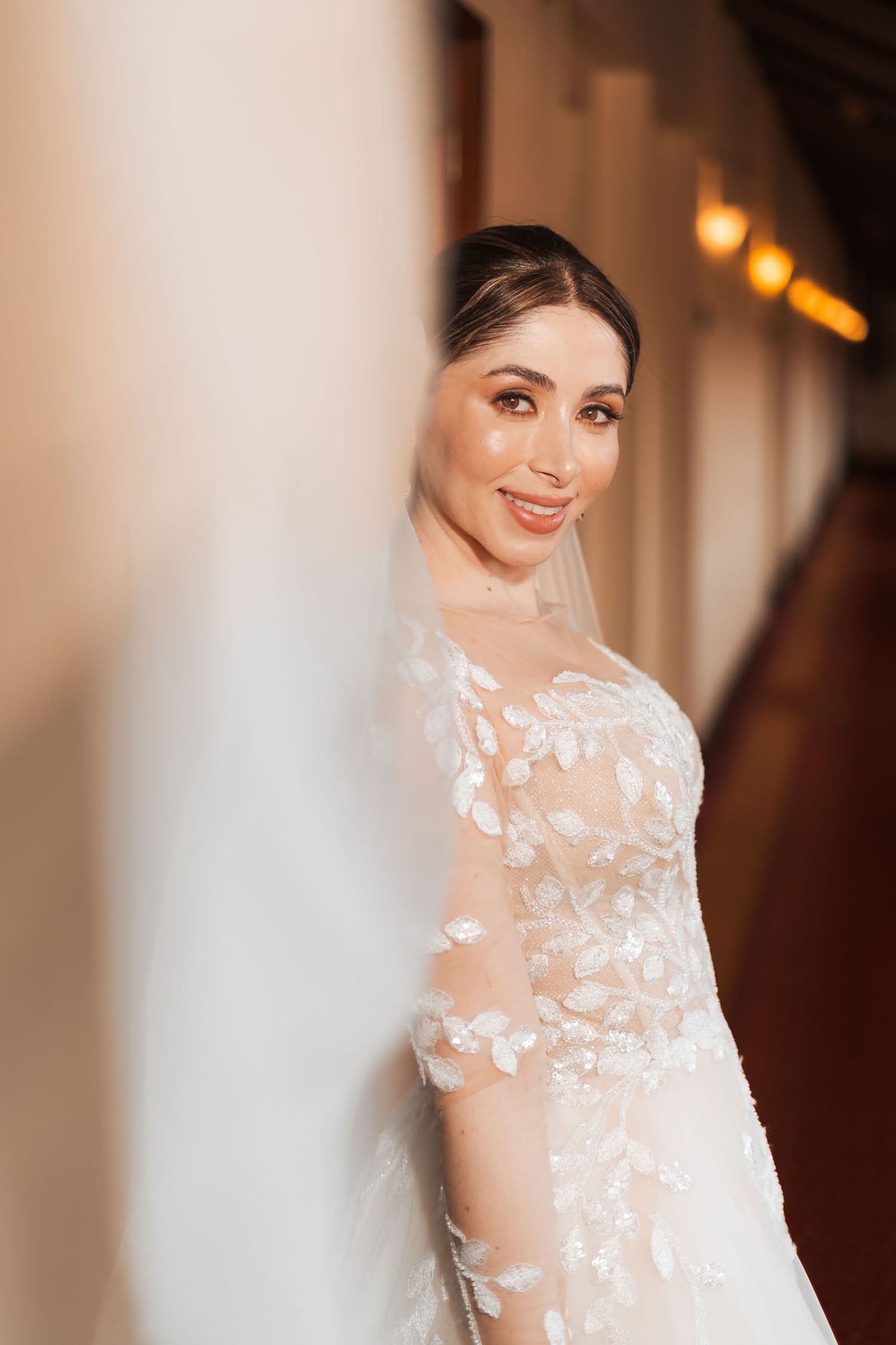 Novia sonriente mira a cámara a través de su velo en primer plano; retrato artístico boda en pasillo interior.