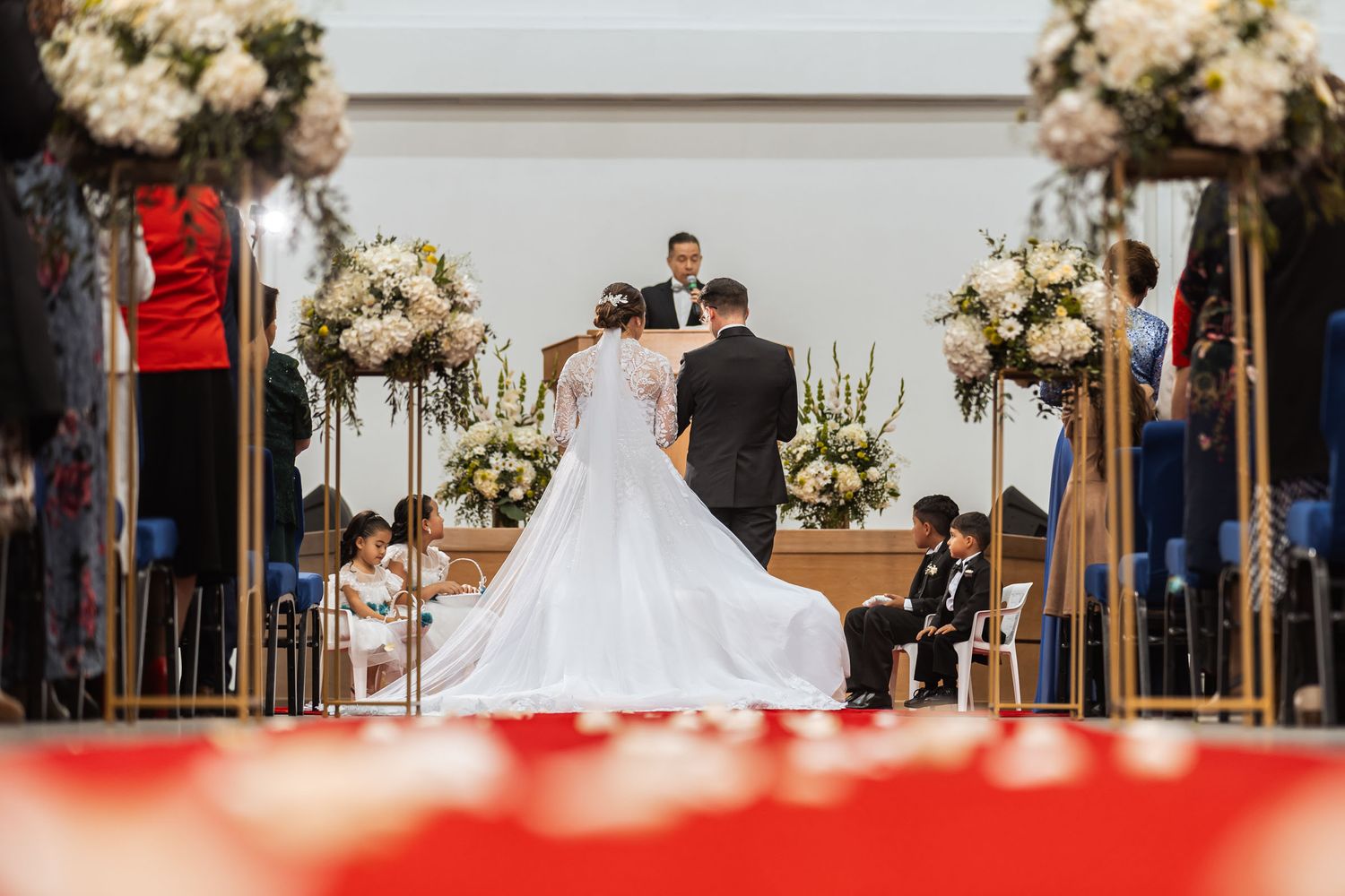 Novios de pie frente al oficiante en el altar durante ceremonia de boda IDMJI, rodeados de flores y público.