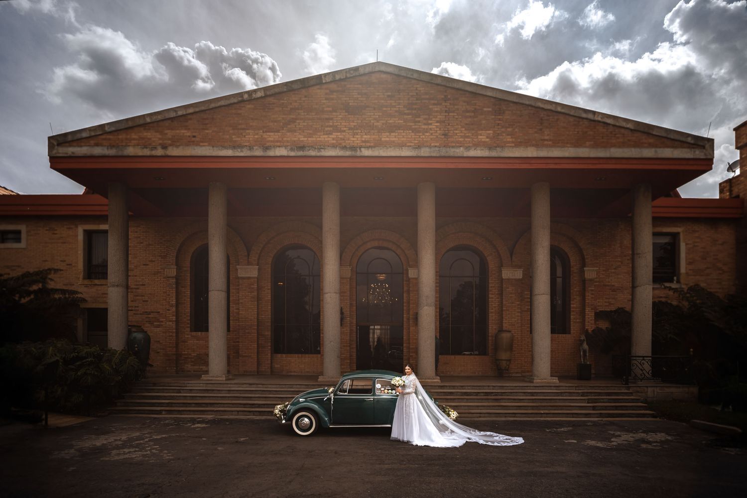 Novia con velo largo y coche clásico VW Escarabajo verde posan frente a imponente edificio con columnas; foto boda.